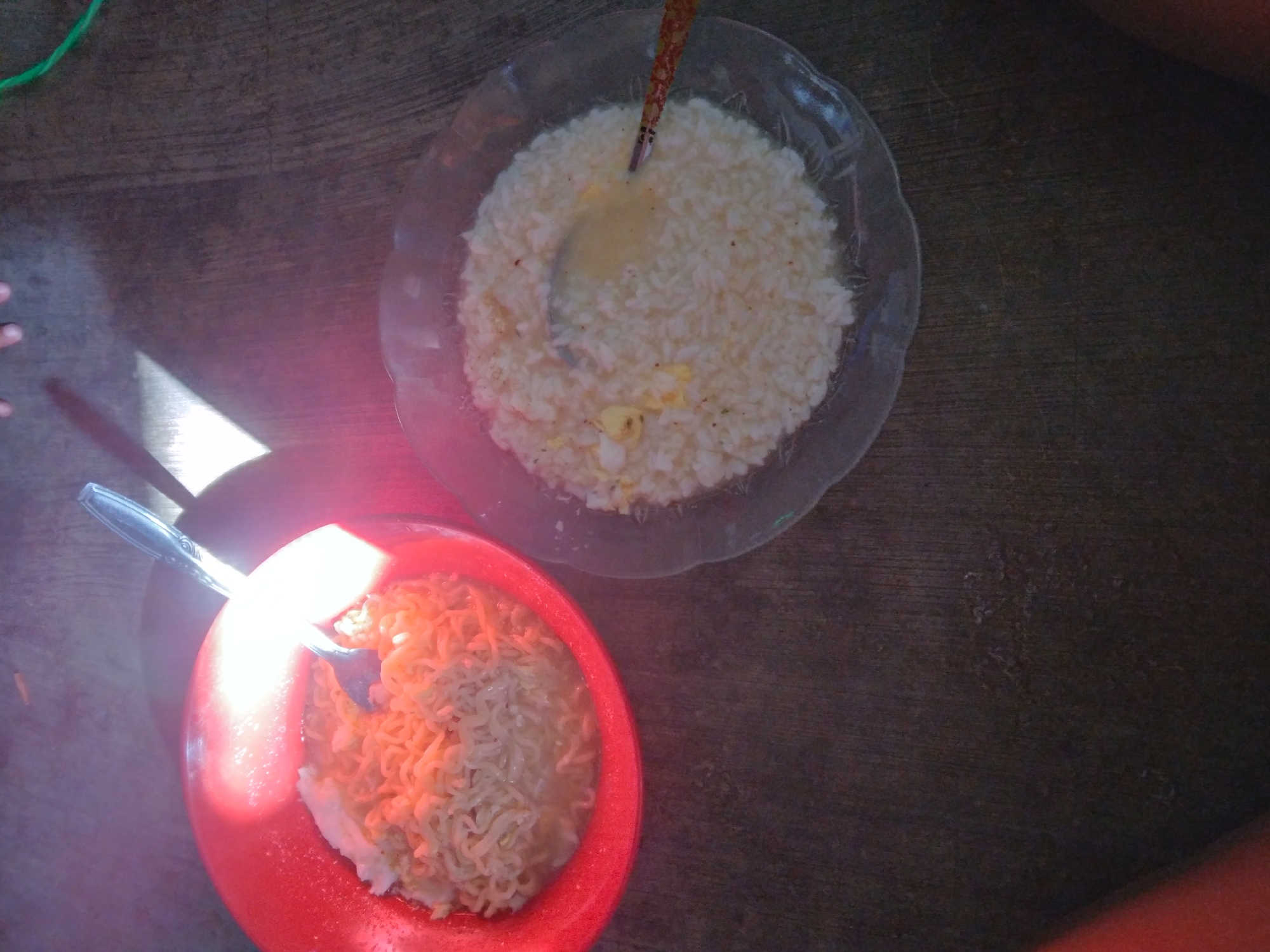 Two bowls of food are placed on a wooden surface. The bowl on the left is red and contains noodles. A silver spoon is in the bowl. The bowl on the right is clear and contains rice. A spoon with a patterned handle is in this bowl. The image appears to be taken in Wringinanom, Indonesia, as suggested by the context. There are shadows present, indicating overhead lighting, possibly from a window or the sun. A green cord is visible in the upper right corner of the image.