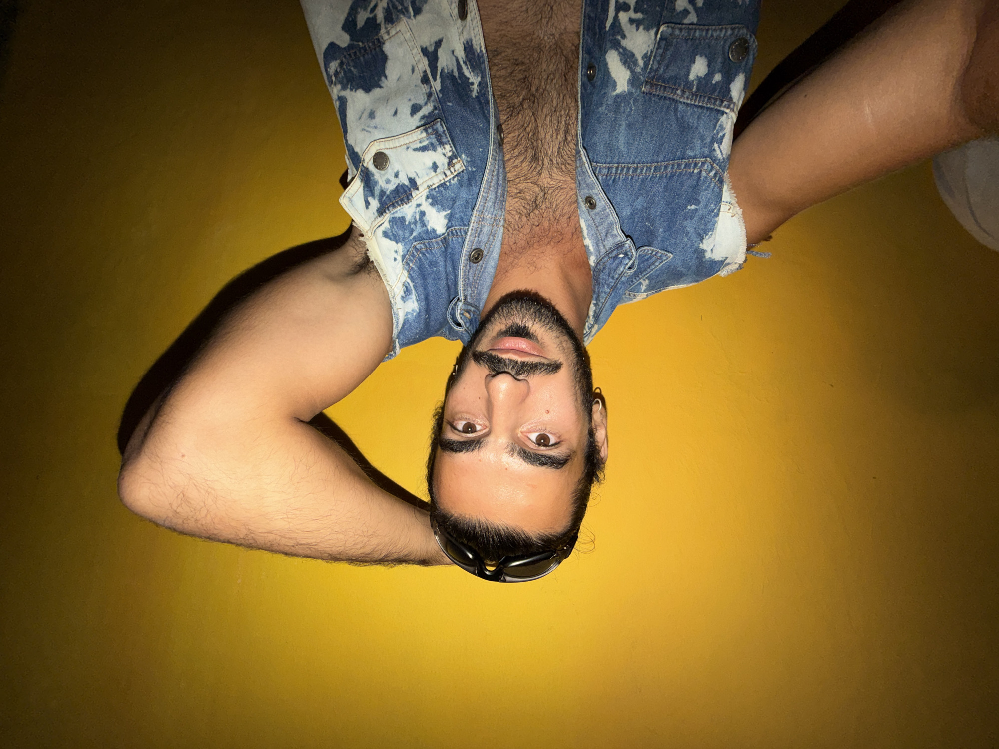 A young man with dark hair, a mustache, and a beard is shown from the chest up, posing against a yellow wall. He is wearing a distressed denim vest with a bleached pattern and a black t-shirt underneath, revealing chest hair. He has sunglasses pushed up on his forehead and is holding his right arm behind his head. The background is a plain, textured yellow wall with uneven lighting. There is no identifiable city, location, or Indonesian context present in the image.