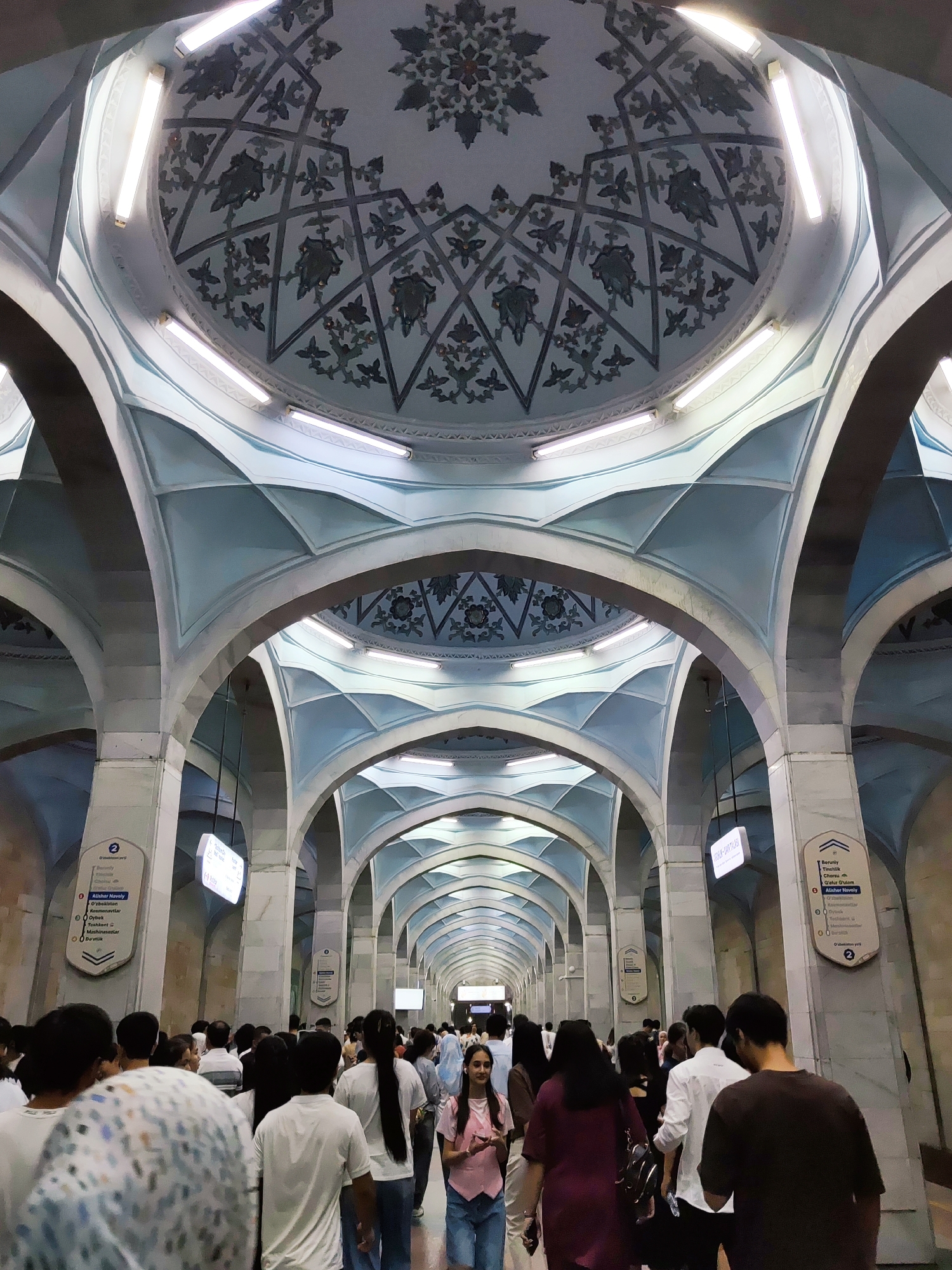 The image depicts the interior of a metro station, likely in Tashkent, Uzbekistan, characterized by its distinctive architecture. The ceiling features a large, ornate dome with intricate geometric patterns and floral motifs in shades of gray and blue. Below the dome, a series of gracefully curved arches creates a long, cavernous passageway. The arches are supported by large, monolithic pillars, and the undersides of the arches are painted in a light blue hue, accentuating the vaulted structure. Fluorescent lights are integrated into the ceiling and along the arches, illuminating the space.
A crowd of people is visible, walking in both directions along the platform. They are dressed in casual attire, with many wearing white shirts. Some individuals are looking at their phones, while others are engaged in conversation. There are informational signs on the pillars, some with text in the Latin alphabet and likely Cyrillic as well, indicating station names and directions. The overall atmosphere is one of a busy public transit hub.