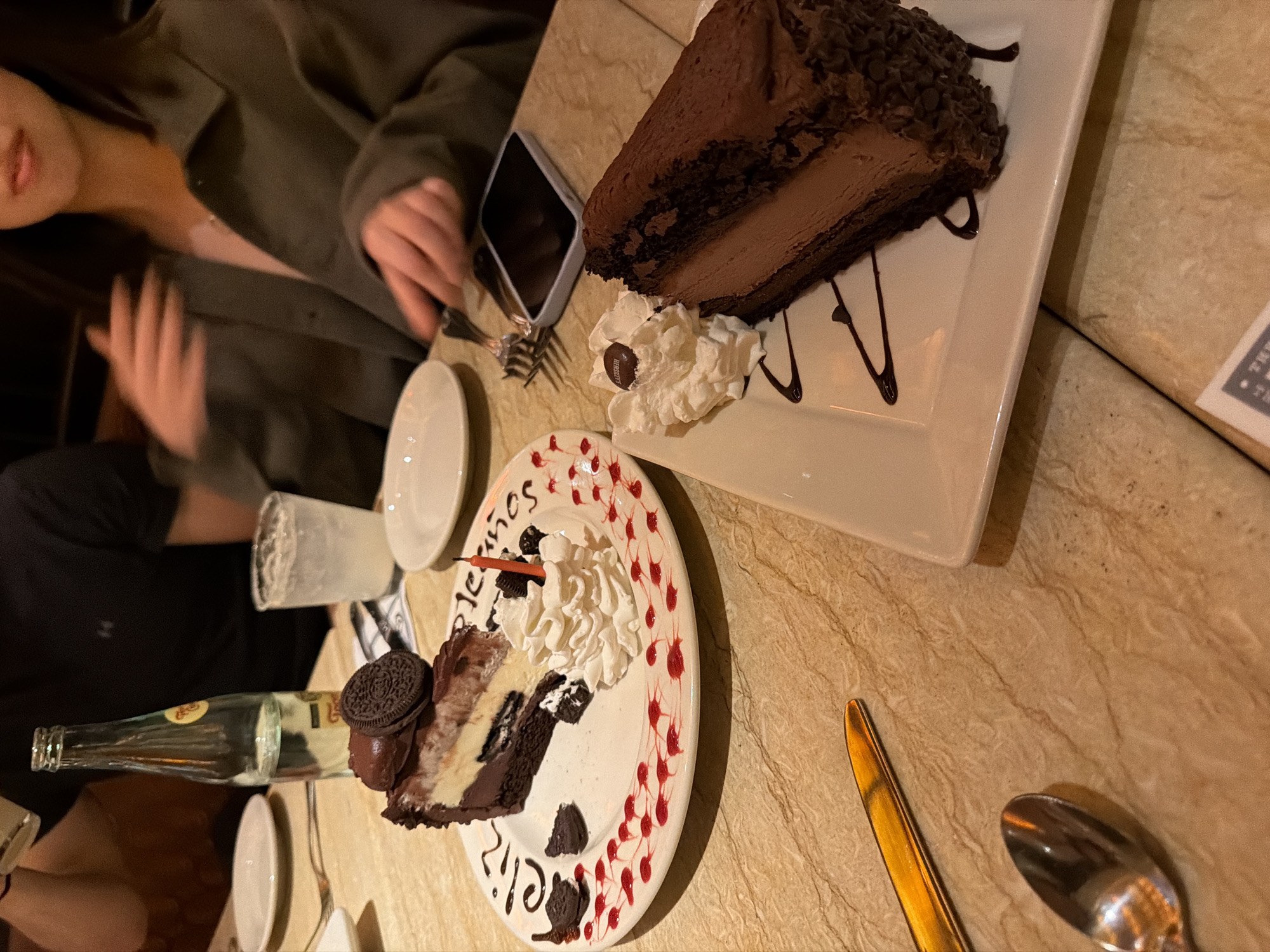 This image depicts a birthday celebration taking place at a restaurant in Puebla, Mexico, during the evening or night, indicated by the warm, artificial lighting. Two individuals are seated at a light brown, marble-patterned table, focusing on two elaborately presented slices of cake.
One slice, a rich dark chocolate cake topped with chocolate shavings, is served on a rectangular white plate with decorative chocolate drizzles. It's accompanied by whipped cream and a Hershey's chocolate kiss. The other slice, appearing to be a cookies-and-cream or Oreo cheesecake, rests on a round white plate. This plate is artfully decorated with "Feliz Cumpleaños" (Happy Birthday) written in chocolate and red decorative dots. A lit birthday candle is inserted into this cake, which is also adorned with whipped cream and an Oreo cookie.
Two people are partially visible. On the left, a person wearing a dark green or grey jacket is seen from the chest up, with part of their smiling face and hands in frame. Another person's arm and torso, dressed in a black shirt, are visible on the lower left.
The table is set with several white plates, forks, a gold-handled knife, a spoon, a clear glass bottle, and a glass containing a clear beverage. A smartphone with a silver edge is placed face down near one of the person's hands. The scene conveys a festive and celebratory atmosphere. Visible text includes "Feliz Cumpleaños" on the round plate and "HERSHEY'S" on the chocolate candy.
