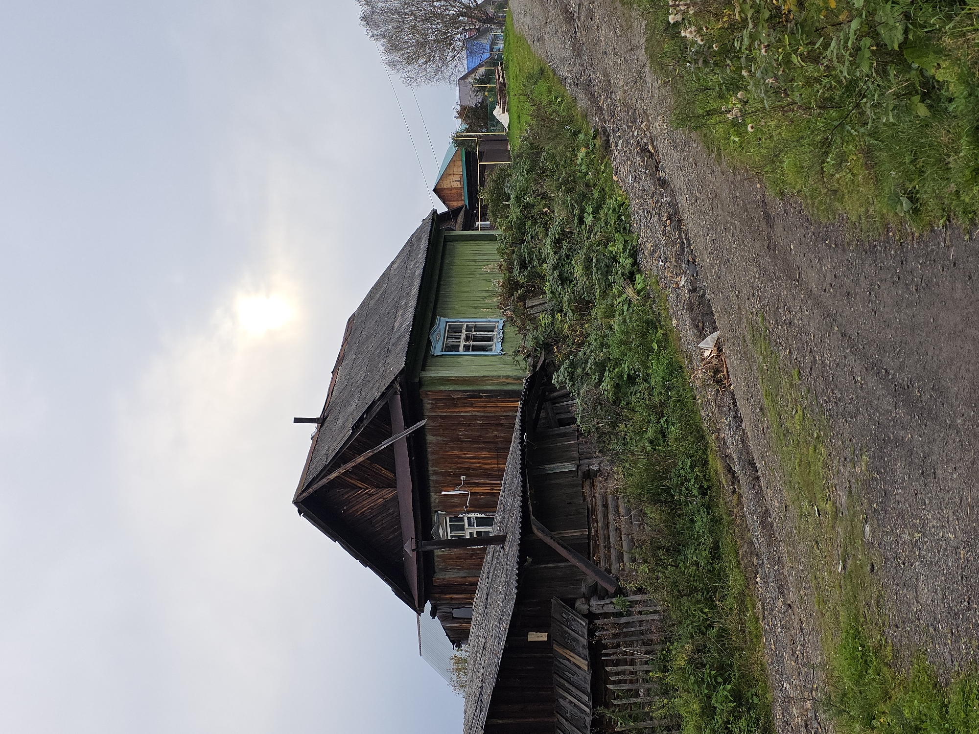 An overcast day in Magnitka, Russia, with a dirt road leading uphill past several weathered wooden houses. The foreground features the rough texture of the road and overgrown vegetation on its right side. The central focus is a two-story wooden house, partially painted green with brown siding, featuring an angled, dark roof. A smaller, darker wooden structure with a corrugated roof is partially visible in the lower left, with a wooden fence in front of it. Behind the main house, other houses with colorful roofs are visible further up the road, along with a bare tree. The sky is a dull grey, with a faint halo around the sun. There are no people or animals visible in the scene.