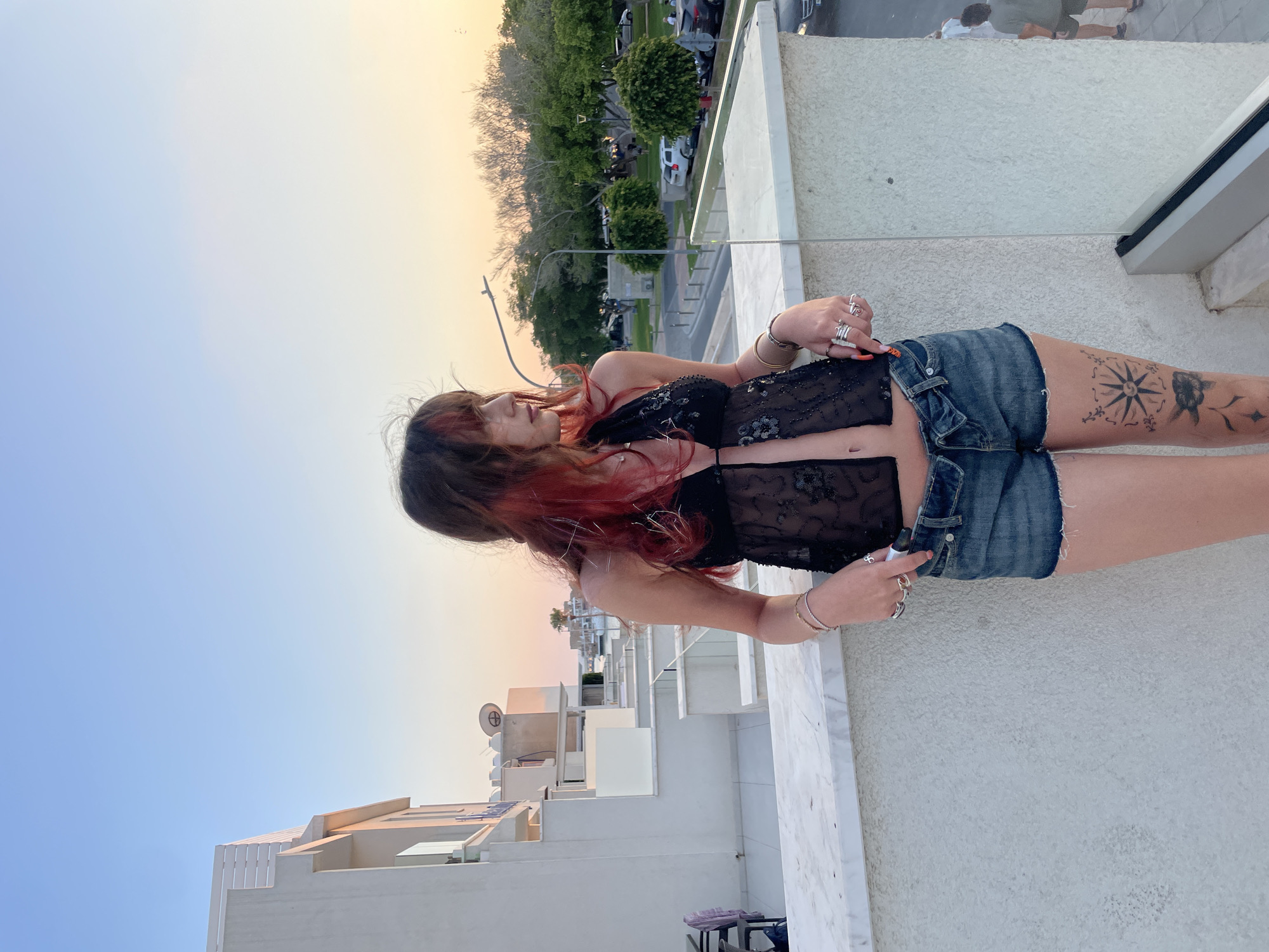 A young woman with dark hair and red streaks stands on a rooftop overlooking a street in Paphos Municipality, Cyprus. She is wearing a black sequined halter top and denim shorts. The sky is a soft blue transitioning to a pale orange at the horizon, suggesting it is either sunrise or sunset. In the background, there are several white buildings with balconies and a street with parked cars and trees. The woman is looking to her right and her left arm is resting on a white marble railing. She has several rings on her fingers and a tattoo on her left thigh.