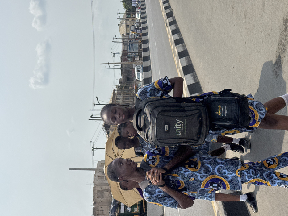 The image captures a group of approximately five to six young individuals, likely students, walking along a paved surface adjacent to a multi-lane road in the city of Ogbaku, Nigeria. The sky is bright with scattered clouds, indicating a daytime scene with direct sunlight casting shadows. All visible individuals wear matching school uniforms featuring a blue, yellow, and white patterned fabric with "SECONDARY SCHOOL OWERRI" inscribed.
In the immediate foreground, a male student on the left wears a dark backpack, has his hands clasped, and looks to his left with a slight smile. To his right, a female student wears a prominent dark grey backpack with a "Unity bank" logo; a smaller black bag labeled "Saints" is attached beneath it. She looks straight ahead with eyes partially closed. Behind these two, other students are visible, one holding a large yellow umbrella overhead, providing shade.
The paved surface they walk on is bordered by a black and white striped curb or barrier, separating it from the vehicle lanes of the road. In the midground and background, multiple vehicles, including white vans, are present on the road. The urban environment features various multi-story buildings, some appearing incomplete or in disrepair, along with utility poles and overhead lines.