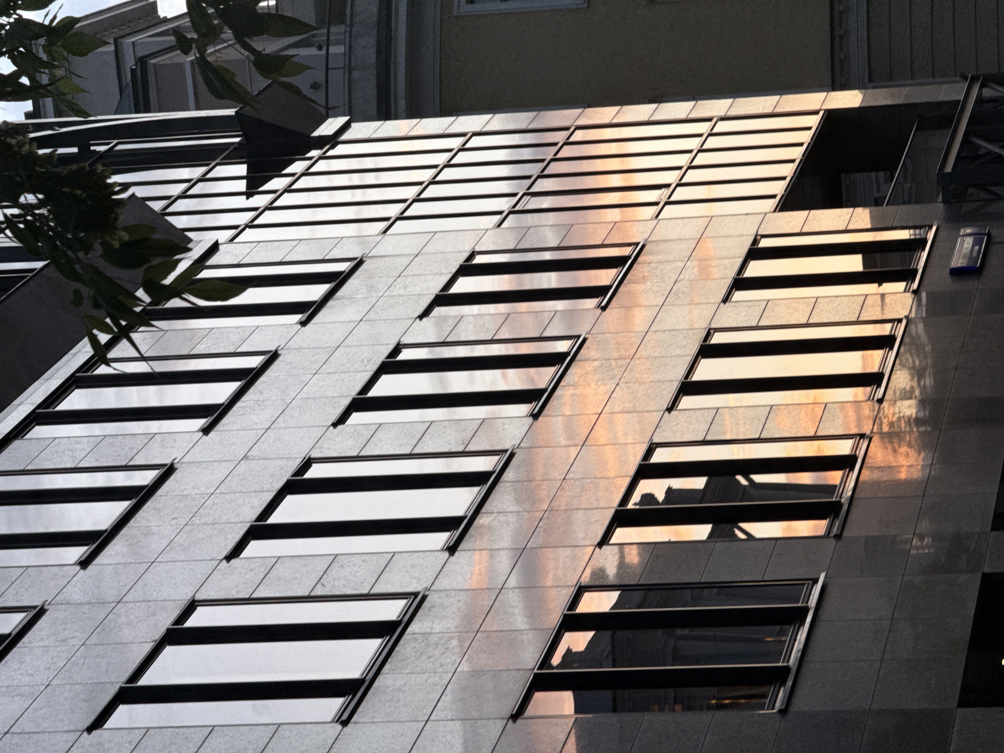 The image depicts the facade of a modern building in Moscow, Russia. The building features a grid-like pattern of square tiles with narrow, horizontal windows set within each tile. The windows reflect the colors of the sunset or sunrise, suggesting it is either early morning or late afternoon. The architecture is contemporary, with clean lines and a somewhat angular design. The building material appears to be a combination of glass and a dark-colored stone or tile. In the top left corner, there are some blurred leaves from a tree, and in the upper portion of the image, a glimpse of neighboring buildings is visible. The building's facade is slanted to the left, and the camera angle is slightly upwards.