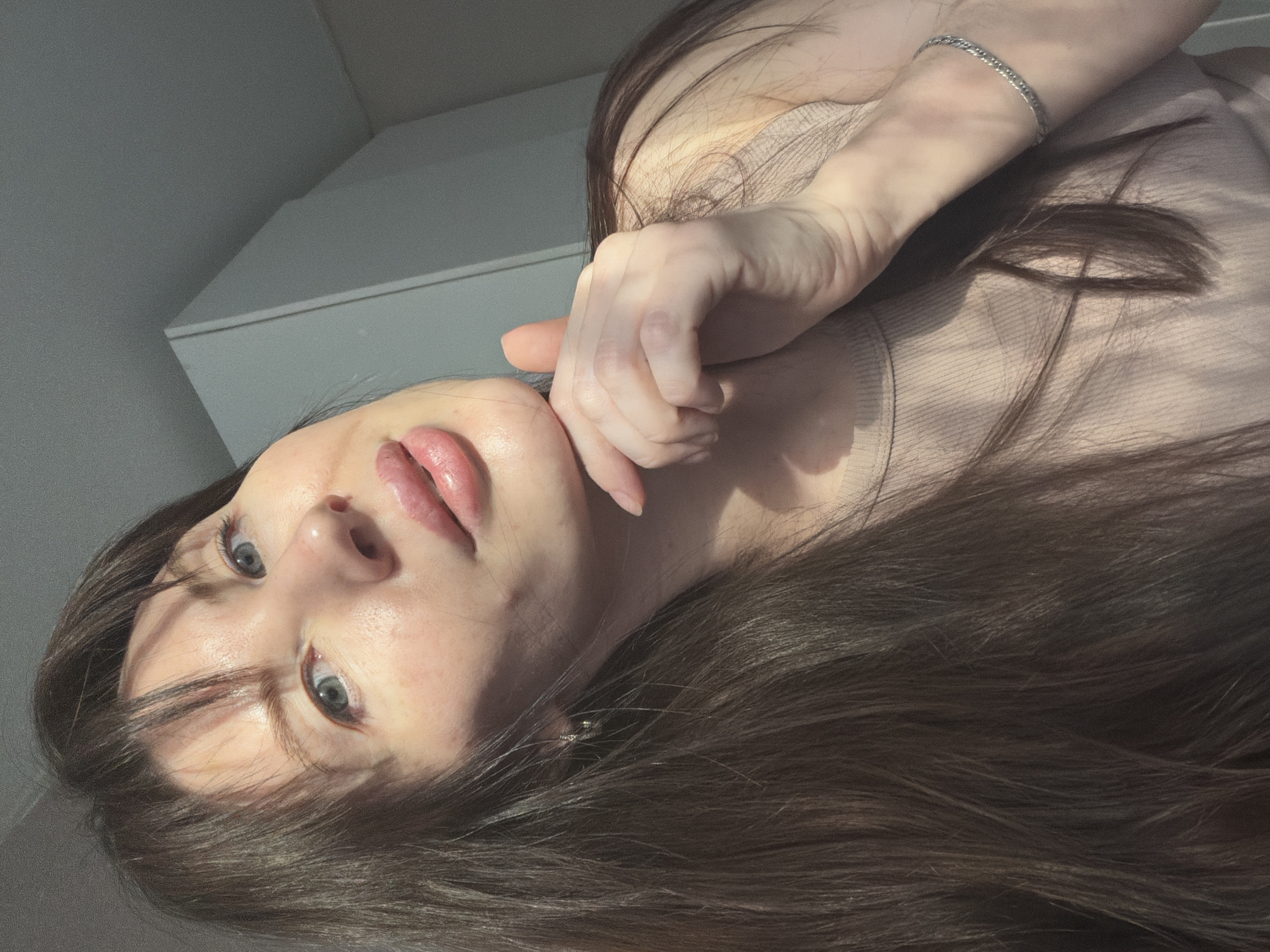 A woman with long brown hair and blue eyes is pictured from a low angle, looking up at the camera. She has fair skin and is wearing a light-colored top. Her right hand is near her face, with her fingers touching her cheek. She wears a silver bracelet on her right wrist. The background is a white wall with shadows. The location is not discernible as Perm, Russia.