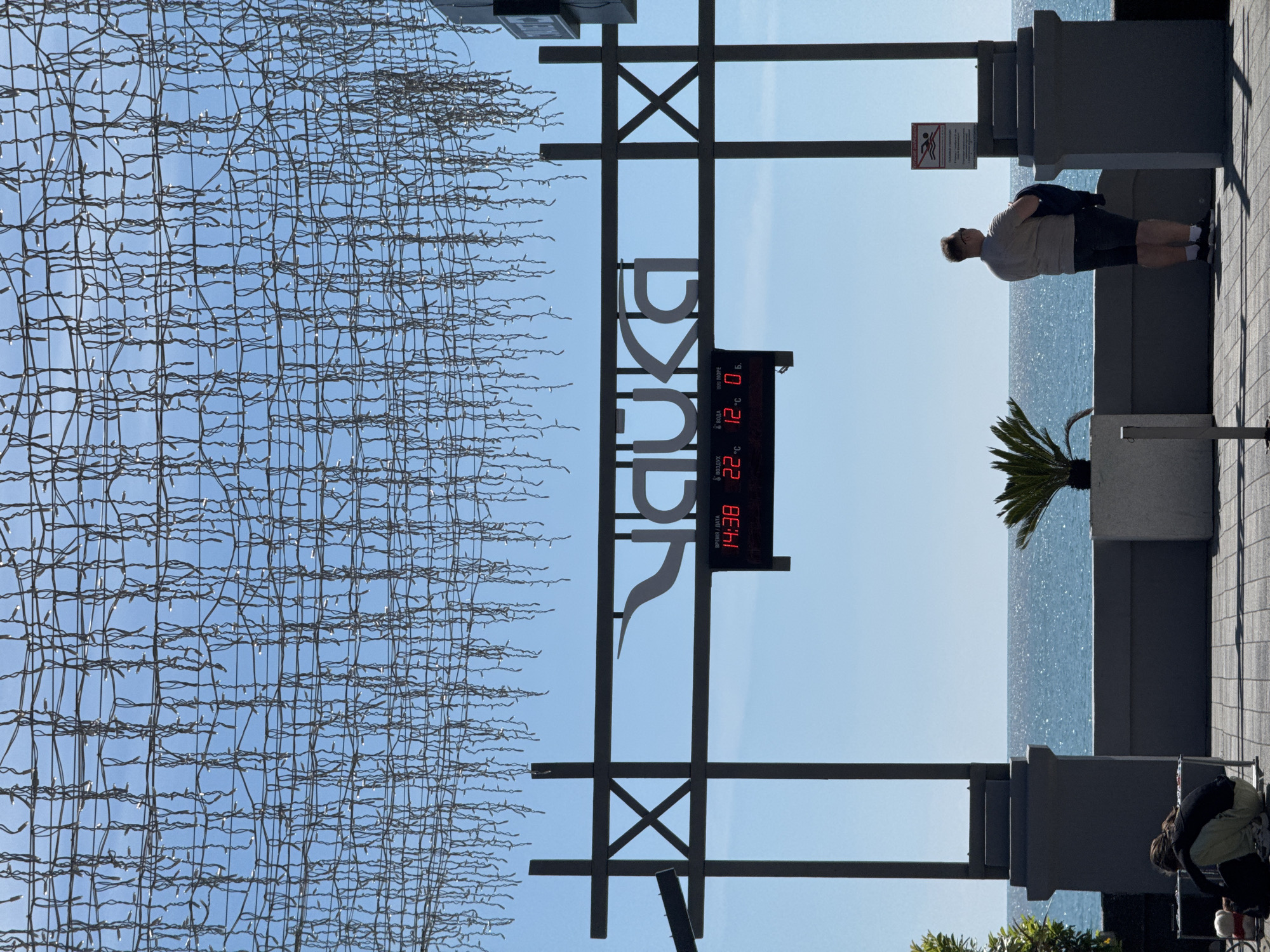 Here is a concise description of the image:
The image depicts the entrance sign to Riviera Park in Sochi, Russia, seen on a sunny day. The sign, rendered in Cyrillic letters, features a digital display showing the time as 14:38, the air temperature as 22 degrees Celsius, and the water temperature as 12 degrees Celsius. Adjacent to the sign is an artificial light structure forming a grid pattern.
Beyond the sign, the scene includes a view of the sea, along with a paved walkway lined with railings. Two individuals are present in the image: one leaning against the railing looking at the sea and another bending down at the base of a railing to the right.