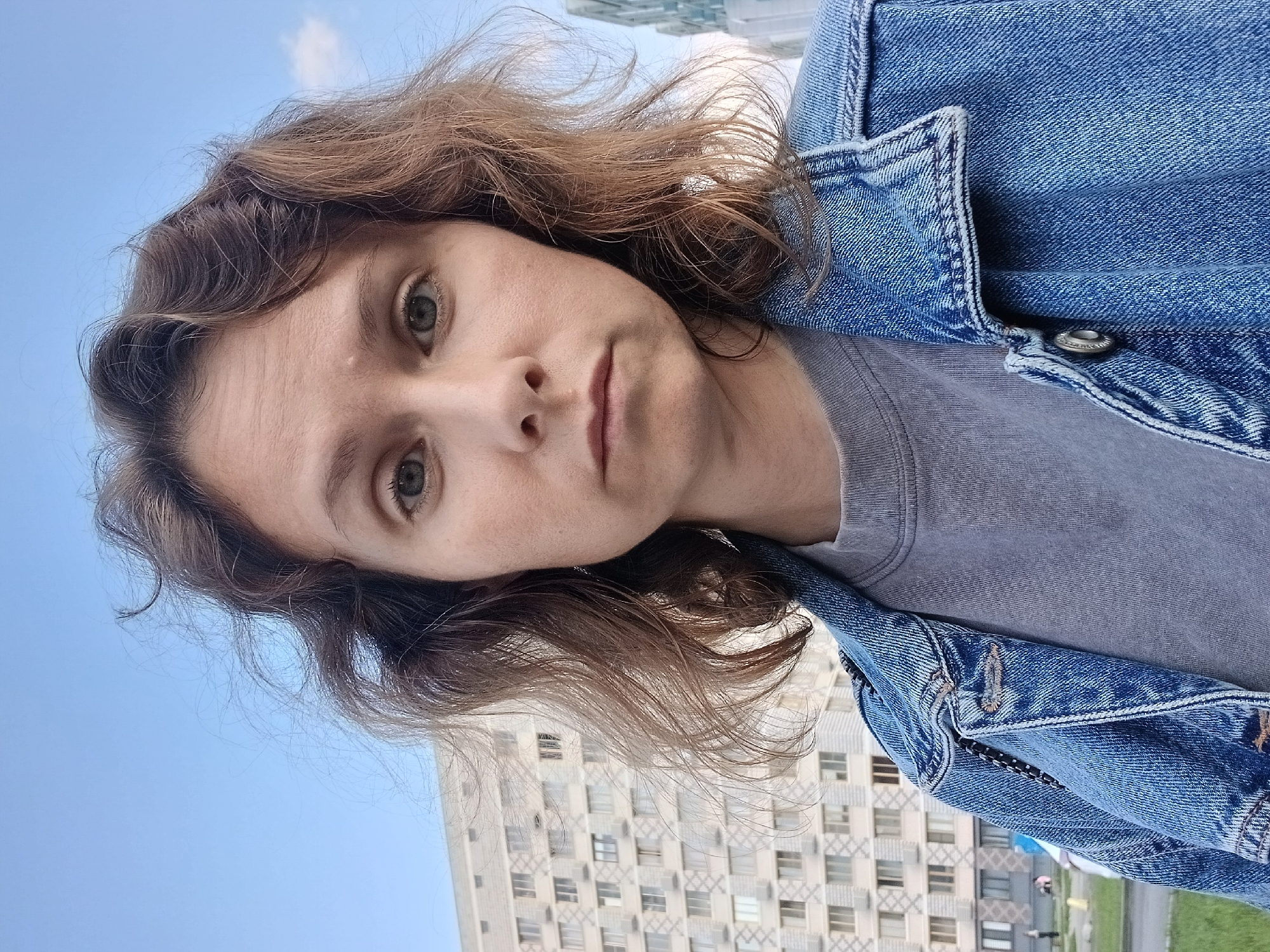 This is a selfie taken outdoors in front of a modern apartment building with numerous windows and balconies in Yekaterinburg, Russia. The sky is clear blue with a few faint clouds. In the foreground, a person with shoulder-length, wavy brown hair is looking directly at the camera. They are wearing a grey t-shirt and a blue denim jacket. Their expression is neutral. The building behind them appears to be a multi-story residential complex. The overall impression is a casual outdoor portrait with an urban backdrop.