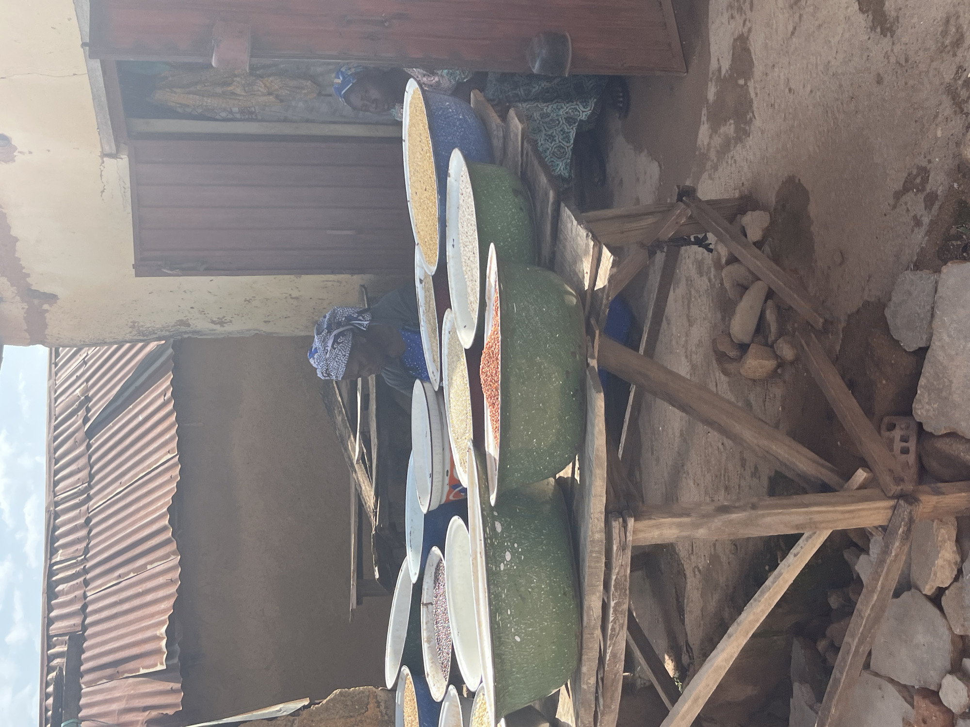 In Jos, Nigeria, a market stall displays an assortment of grains in large metal basins. Two women are present, one tending to the grains and another seated in the background. The stall is constructed from rough-hewn wood and is covered by a corrugated metal roof. The surrounding environment appears to be dusty and undeveloped.
