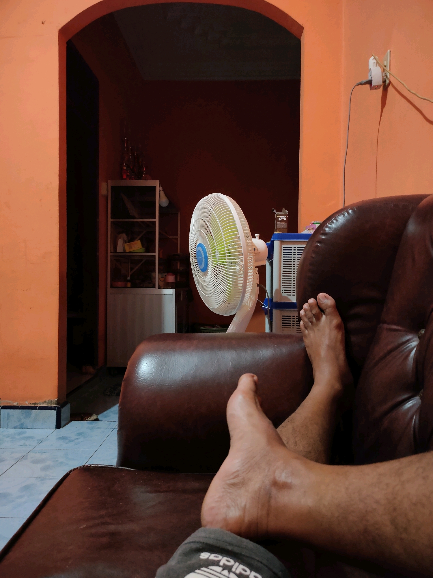 A first-person perspective shot shows a pair of bare feet resting on a dark brown leather couch in what appears to be a living room. The feet are positioned in the lower right quadrant of the frame, with the ankles and lower legs visible. The couch has a glossy sheen, reflecting ambient light.
In the background, an orange wall dominates the scene. To the left, an arched doorway leads to a darker interior space. A white oscillating fan is visible in the mid-ground, positioned to the left of center. Behind the fan, a white and blue two-tiered appliance, possibly a cooler or storage unit, is present. Further back, a metal shelving unit holds various items, including what looks like bottles or containers. The floor is tiled with a pattern of light blue squares. A white electrical outlet with a plug and cord is mounted on the orange wall to the right.
The scene suggests a domestic setting, likely a home in Punggulan, Indonesia, during the evening or night, given the artificial lighting. There is no indication of any specific event or interaction occurring. The focus is on the resting feet and the immediate environment.