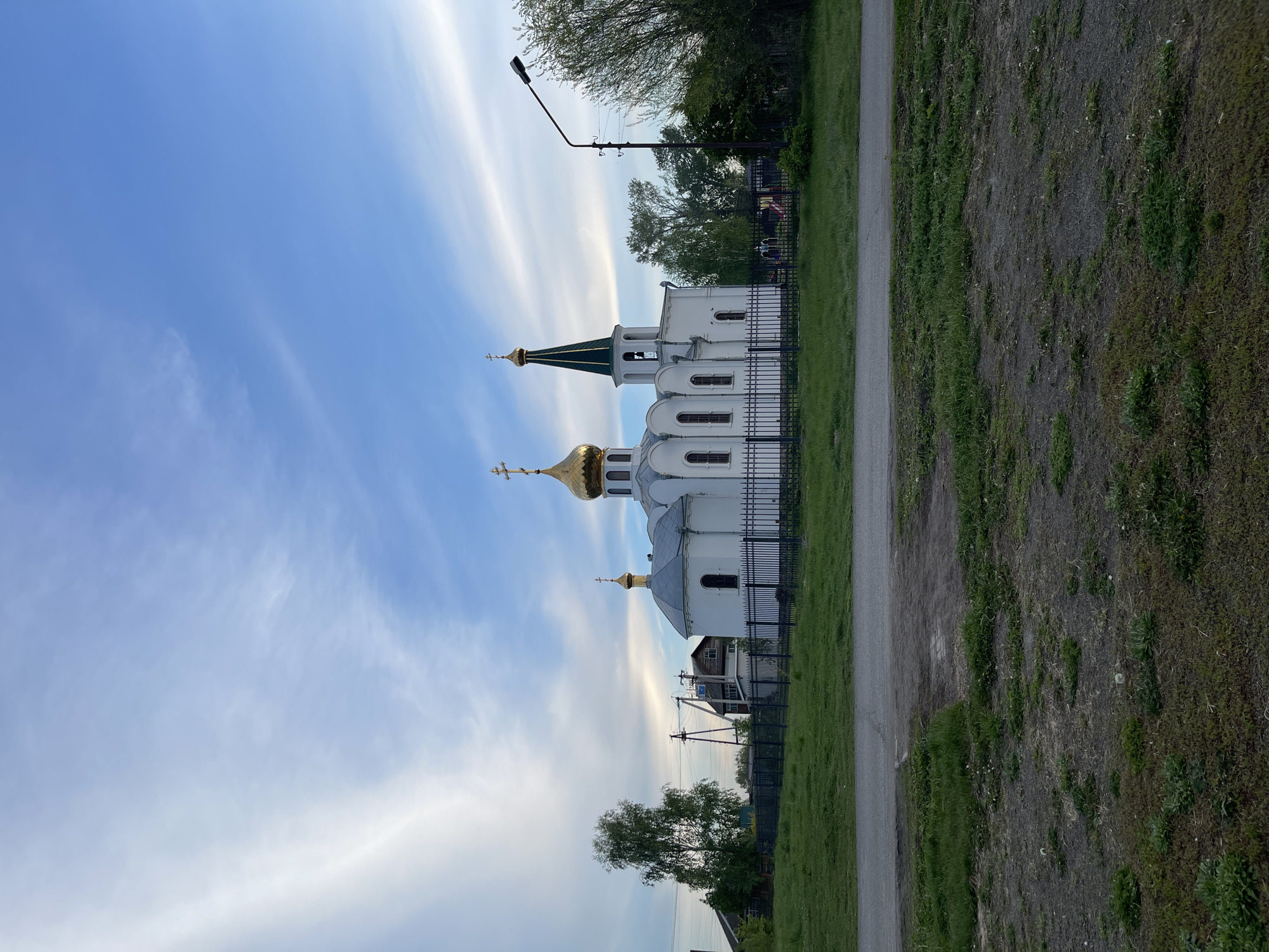 A landscape photograph shows a white, Orthodox church with golden domes in Verkh-Katunskoye, Russia. The church is located behind a dark metal fence, bordering a grassy area and a narrow paved path. A streetlight stands near the church. The sky is blue with wispy white clouds. In the foreground, some trees and parts of houses are seen.