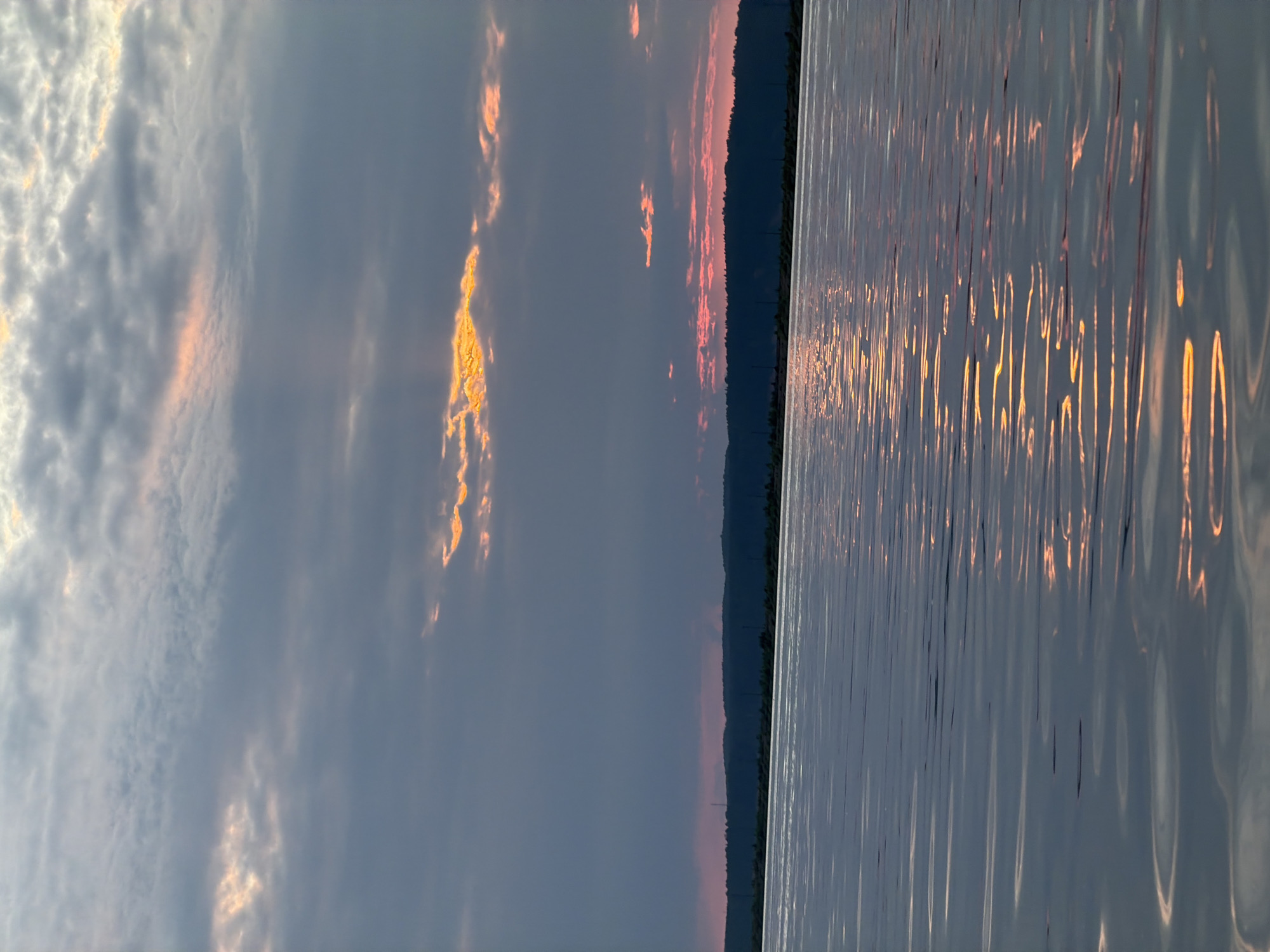 The image depicts a serene natural landscape at dusk, likely on the Kama River in Chaikovsky, Russia. The sky is filled with dramatic clouds, some illuminated with hues of orange and pink from the setting sun, while others are a deep grey. Rays of light pierce through the clouds, creating a striking visual effect. Below the sky, a dark, silhouetted treeline stretches across the horizon, suggesting a forested embankment. The foreground is dominated by the tranquil surface of the river, its waters reflecting the colors of the sky and the muted light, creating a shimmering, textured effect. The overall atmosphere is one of peaceful twilight in a natural, undeveloped setting.