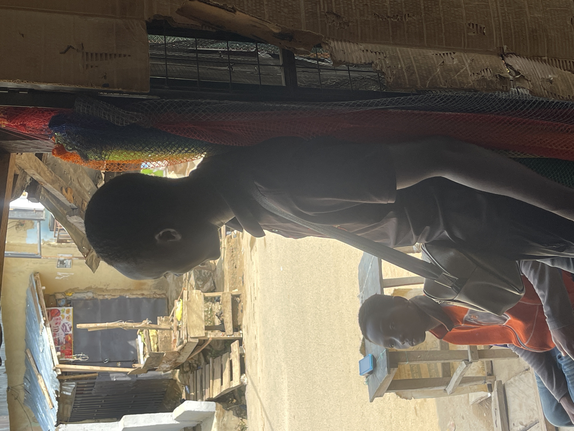 The image shows two young men standing in an urban setting likely within Abuja, Nigeria. The environment consists of dilapidated structures with exposed wooden planks, corrugated iron sheets, and concrete elements. One of the men is wearing a light-colored shirt and carrying a bag over his shoulder. The second man is in an orange and white shirt with jeans. Above them, there is an array of colorful mesh-like materials attached to a damaged ceiling. The background includes hints of signage on a building and general street clutter.