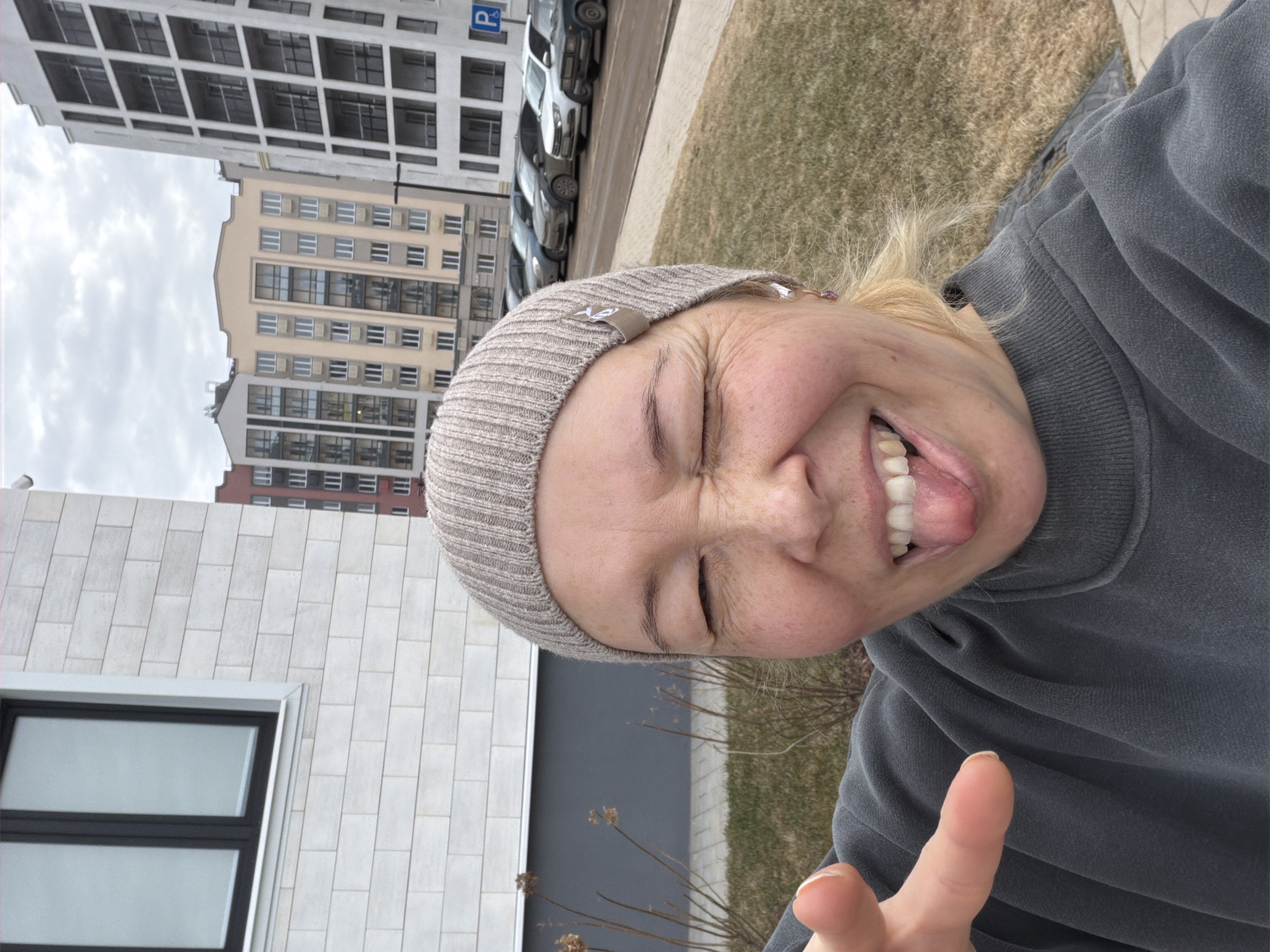 A light-skinned person with blonde hair is taking a selfie outdoors. They are wearing a gray knit cap and a gray sweatshirt. Their eyes are closed, and they are sticking their tongue out while making a finger gesture toward the camera.
Behind them, there's a mix of modern buildings. One building is white with large windows, and another is a tan high-rise. Cars are parked along the side of the street. The ground appears to be covered with dormant grass.
The location is Pervomaysky, Russia, as implied by the prompt.