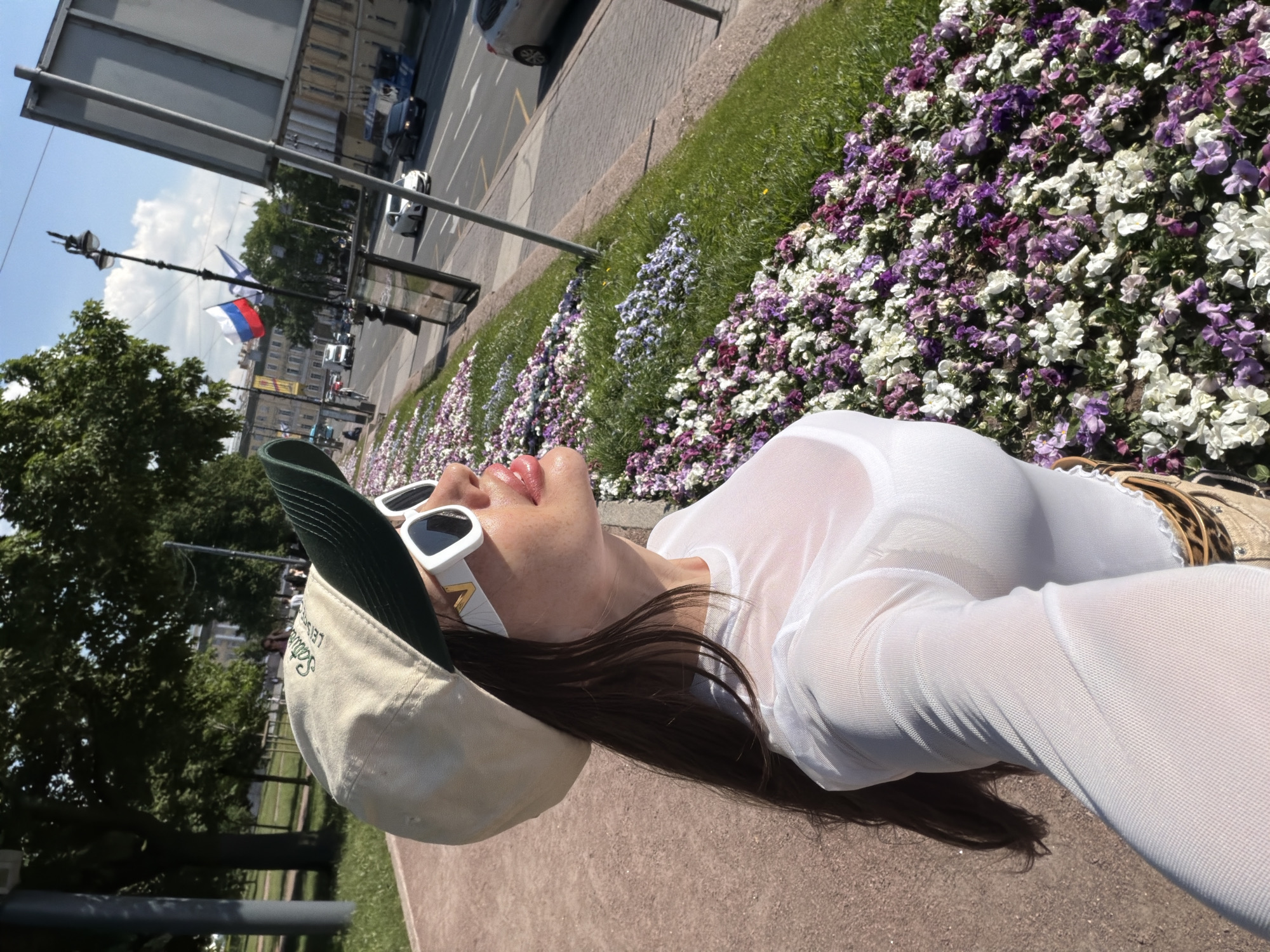 A woman is standing on a pedestrian walkway next to a flowerbed filled with purple and white flowers. She is wearing a white sheer long-sleeved shirt, a beige baseball cap with a dark green brim, and white sunglasses. The flowerbed runs alongside a street where cars are driving, and buildings are visible in the background. Russian flags are flying on flagpoles in the distance. The scene appears to be in Saint Petersburg, Russia, likely near a park or boulevard given the presence of trees and a well-maintained sidewalk. The lighting suggests it is daytime with clear skies.