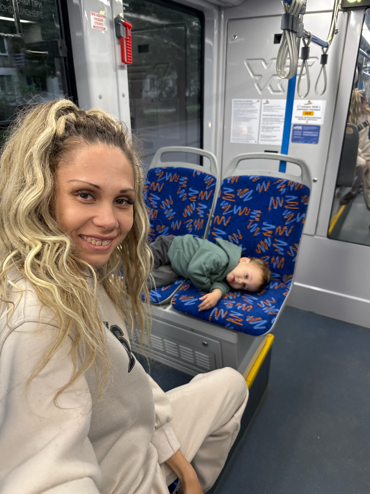 A woman with blonde hair pulled into a high ponytail smiles at the camera, her head and shoulders filling the left side of the frame. She is wearing a cream-colored hooded sweatshirt. To her right, a young child in a green hooded sweatshirt is lying on a blue seat with an orange and white pattern. The child is looking away from the camera. The setting appears to be the interior of a public transport vehicle, likely a bus or train, with grey walls and blue seats visible. There are handrails and signage in the background. The overall lighting is natural, suggesting it is daytime.