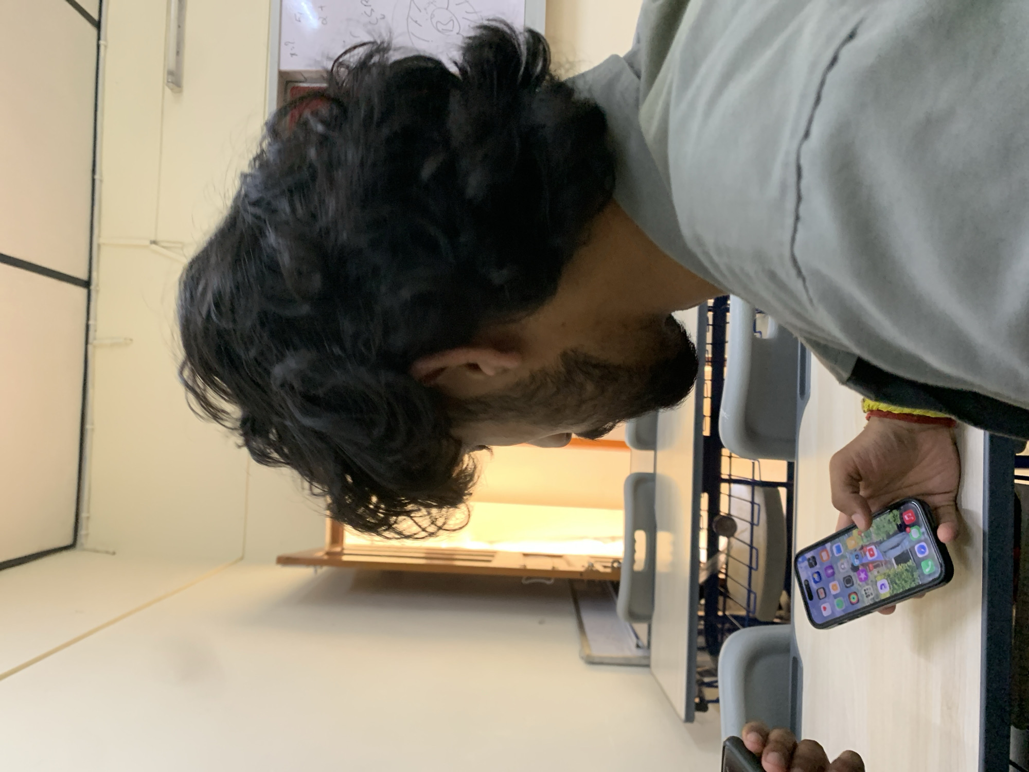 A man with dark, curly hair and a beard is seated at a table, looking at his phone. The phone displays a grid of app icons, with a picture of a person in denim jeans as the wallpaper. The man is wearing a grey shirt and has a red and yellow bracelet on his wrist. In the background, there are empty desks and chairs, suggesting a classroom or meeting room. The lighting is bright, and there's a whiteboard visible on the wall with some writing. The overall setting appears to be in Dharamshala, India, likely an educational institution or administrative office.