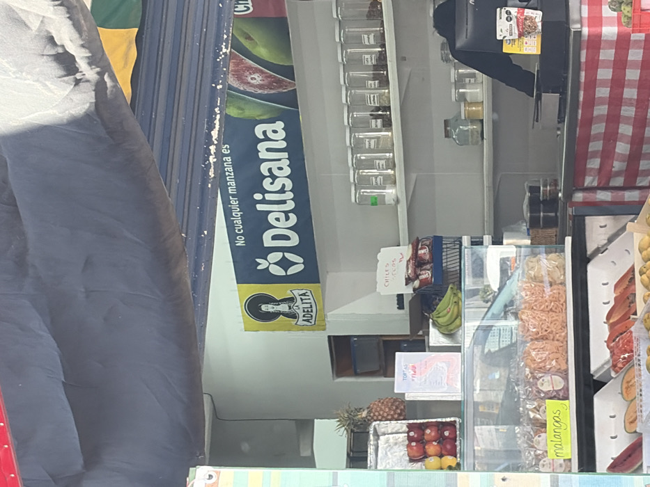 A market stall with produce including apples, pineapples, bananas, and papaya. Jars line a shelf, with packages of chiles nearby. A sign advertises "Delisana," stating, "No cualquier manzana es". The stall has a red and white checkered covering. Transparent containers display mangoes.