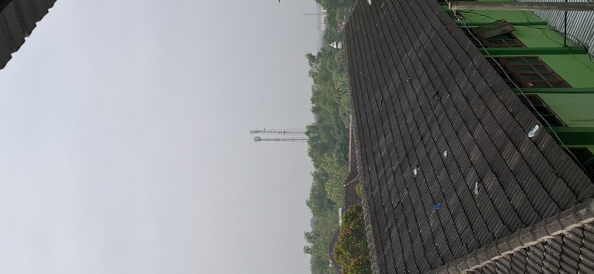 The image depicts an overcast sky above a landscape of rooftops and trees. In the foreground, a dark, corrugated tile roof dominates the lower half of the frame, showing signs of wear and tear with scattered debris, including what appears to be a blue object, possibly a child's toy, and several white plastic items. Beyond this roof, a second, red-tiled roof is visible, partially obscured by dense green foliage. In the middle ground, a line of lush green trees stretches across the horizon, interspersed with buildings. Further in the distance, two prominent cellular towers stand tall against the hazy sky. A small, white dome structure, potentially part of a mosque, is visible among the trees on the right side of the image. The overall scene suggests a semi-urban or suburban environment under a subdued, possibly humid, atmosphere. The location is identified as Wonorejo, Indonesia.
