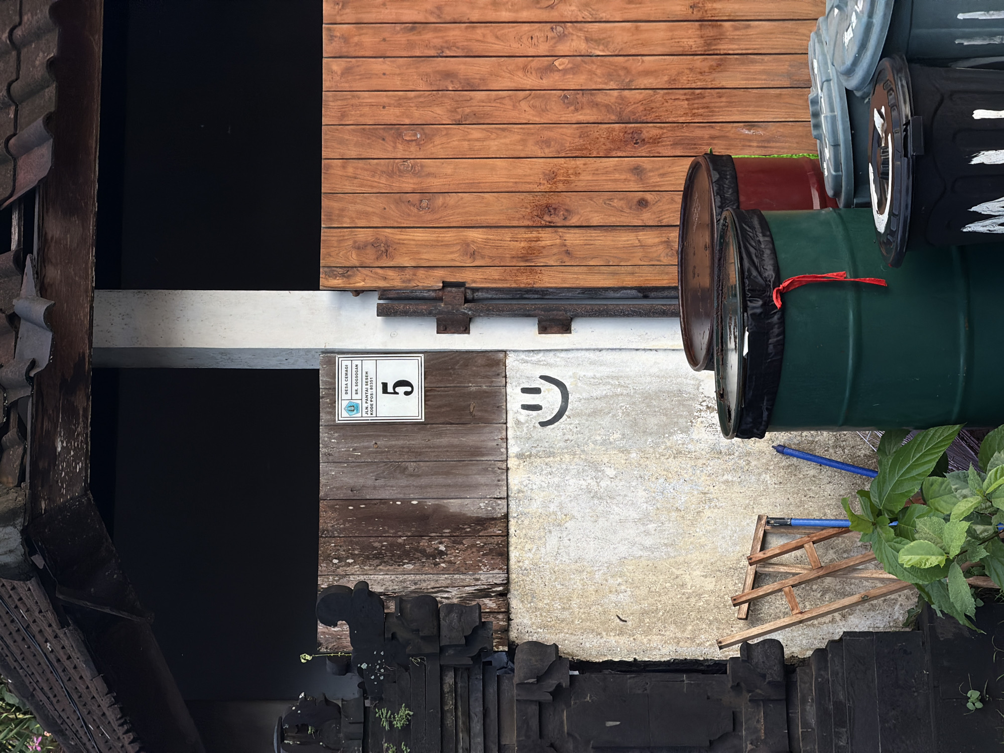 Captured from a high angle, the image presents a view from above, encompassing various elements. The scene includes a building structure with a brown wooden exterior and roofing. A sign displaying the number "5" is affixed to a wooden surface. Adjacent to this, a smiling face has been drawn. To the right of these are several barrels, stacked vertically. The ground beneath features a concrete surface littered with a small wooden structure and a blue pipe. Green plants are positioned near the lower right corner of the frame. The text on the sign indicates that this scene is located in Desa Cemagi, BR. Sogsogan, in Mengwi, Indonesia, specifically on Jalan Pantai Seseh. The postcode is indicated as 80351.