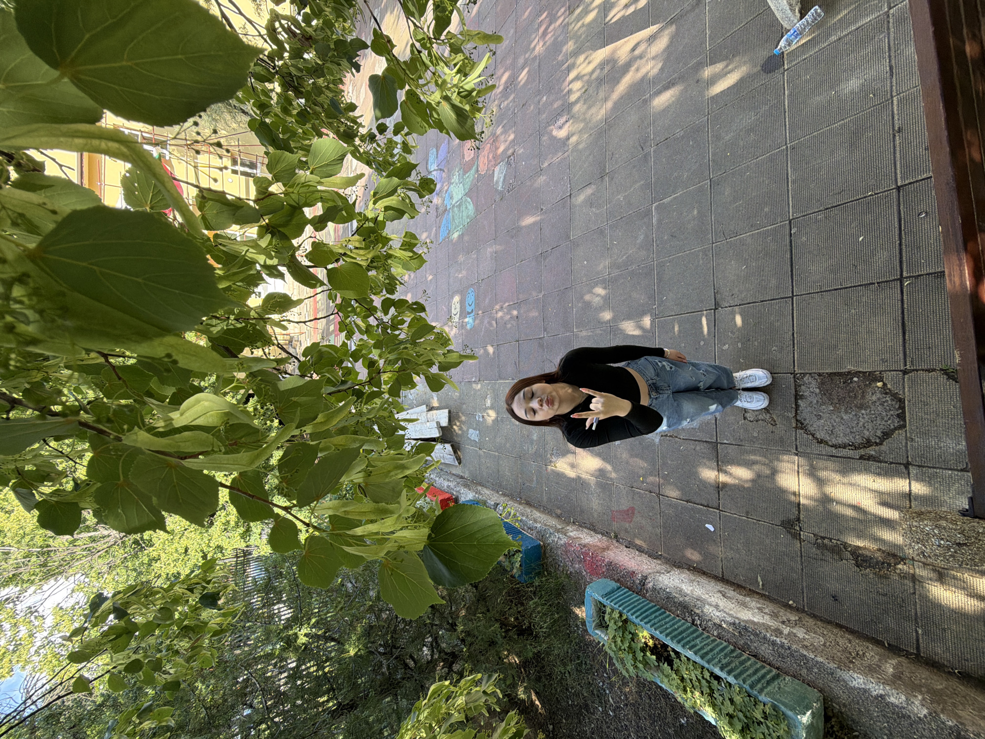 An outdoor, high-angle shot, taken from an elevated position looking down, captures a young woman posing on a paved ground, framed by lush green tree foliage. The image is rotated, with the woman appearing to look upwards towards the camera.
The primary subject is a young woman with dark hair, wearing a black long-sleeved top, light blue ripped jeans, and white sneakers. She stands with a playful expression, making a "rock on" hand gesture towards the camera.
The setting appears to be a public outdoor space, possibly a courtyard or pedestrian area, with a distinctive paved ground made of square, textured concrete tiles. Scattered across the ground are colorful chalk drawings, including smiling face characters and abstract designs. Strong sunlight casts dappled shadows of tree leaves and branches across the ground, indicating a bright, sunny day.
To the right, a dark brown wooden railing or edge of a structure is visible. Along the lower edge of the paved area, there are green, ribbed planters containing small plants. A clear plastic water bottle, identified by legible text as "BANKYA" brand, lies on the ground near the woman's feet. In the background, visible through the surrounding tree leaves, a light yellow building with scaffolding can be seen, suggesting nearby construction or renovation. The scene depicts a casual moment in Bobov Dol, Bulgaria.