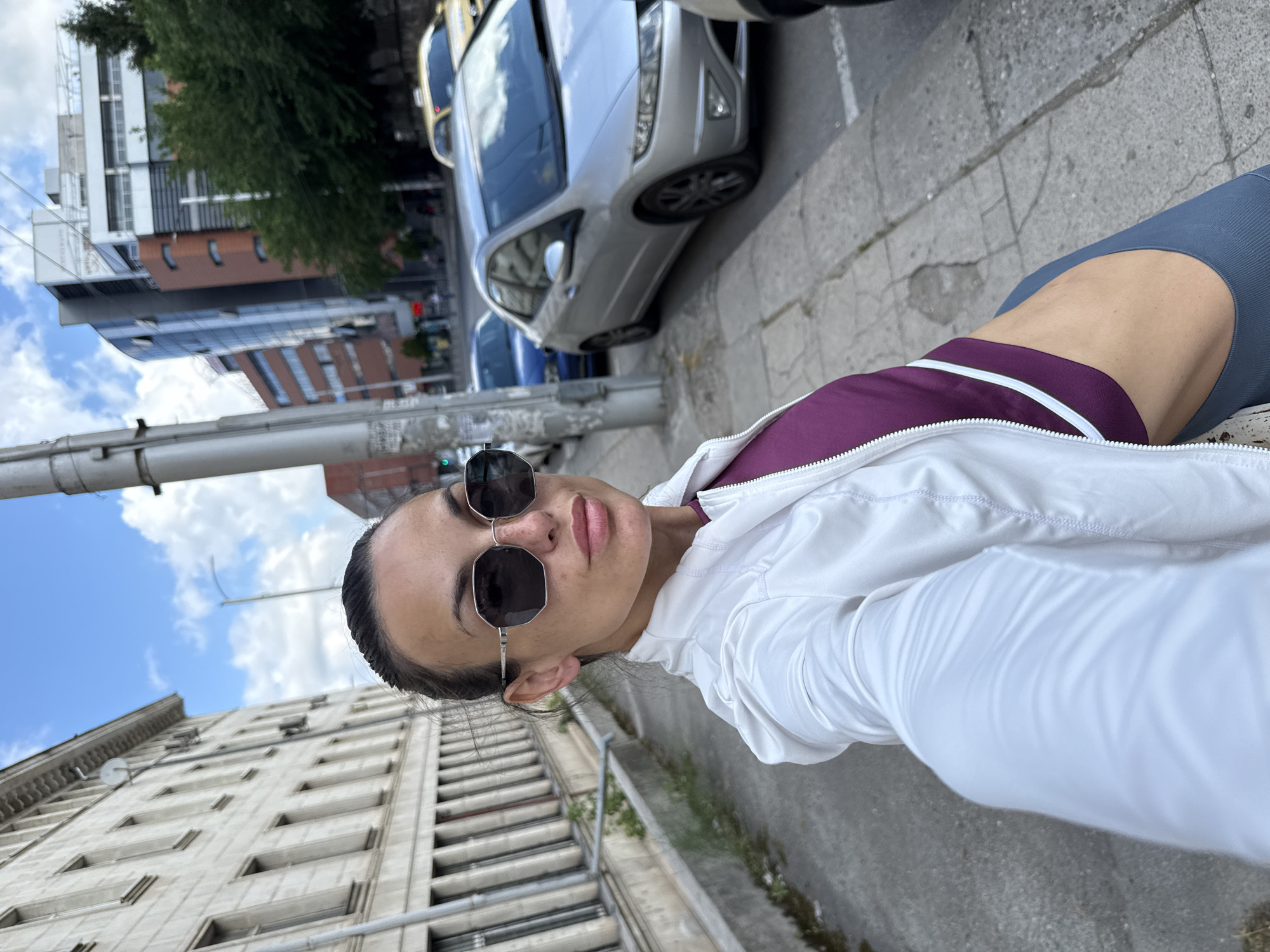 A woman is taking a selfie on a city sidewalk in Pleven, Bulgaria. She is wearing a white jacket over a purple athletic top and gray leggings. She has dark hair pulled back, and is wearing large, dark sunglasses. Behind her, on the left, is a large, light-colored building with many windows and ornate detailing. On the right, there is a sidewalk, a pole, a silver car, and a yellow taxi cab. Further in the background, modern buildings with glass facades and trees are visible. The sky is blue with scattered clouds.