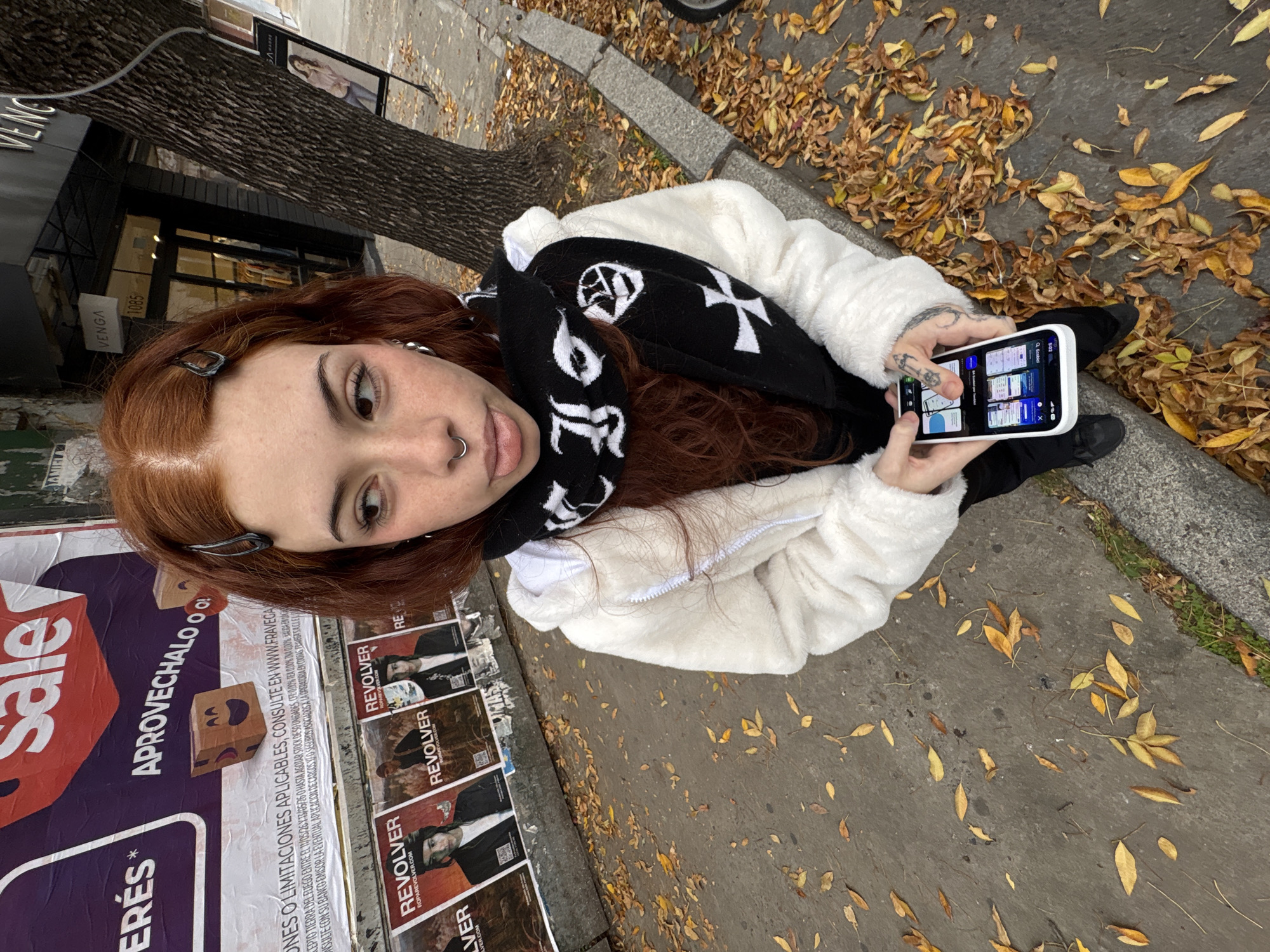 A woman with red hair, a nose ring, and barrettes is taking a picture with her phone on a street in Buenos Aires, Argentina. She is wearing a white fuzzy jacket and a black scarf with white gothic lettering. The street is littered with fallen leaves. Behind her is a wall covered with posters, including ones advertising "Sale" and the clothing brand "Revolver." A store called "Venga" is visible with the number 1085 on its awning. The woman is standing near a curb.