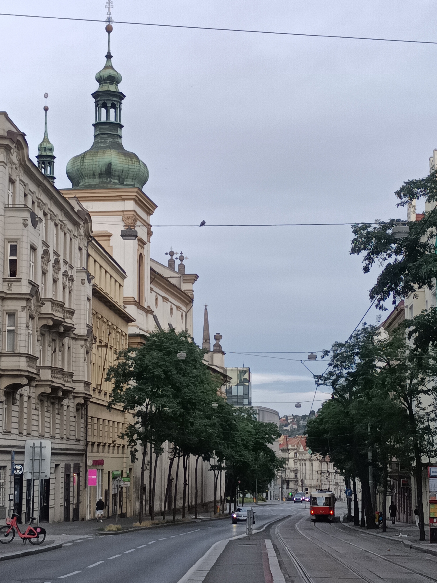 The image depicts a street scene in Prague, Czechia, likely in the Vinohrady district, characterized by its historic architecture and tram system. In the foreground, a red tram with its lights on is traveling away from the viewer on tram tracks. The street curves to the right. On the left side of the street, a row of ornate, multi-story buildings with balconies and detailed facades line the road. A red bicycle is parked on the sidewalk. On the right side of the street, more trees and buildings are visible, with a pedestrian walking away from the viewer.
Dominating the left side of the image are the distinct green onion domes of a church or cathedral, topped with crosses. The sky is overcast and grey. Overhead, electrical wires for the tram system are strung across the street. In the distance, more buildings are visible, with hills and cityscape beyond. The overall atmosphere is that of a European city street on a cloudy day.