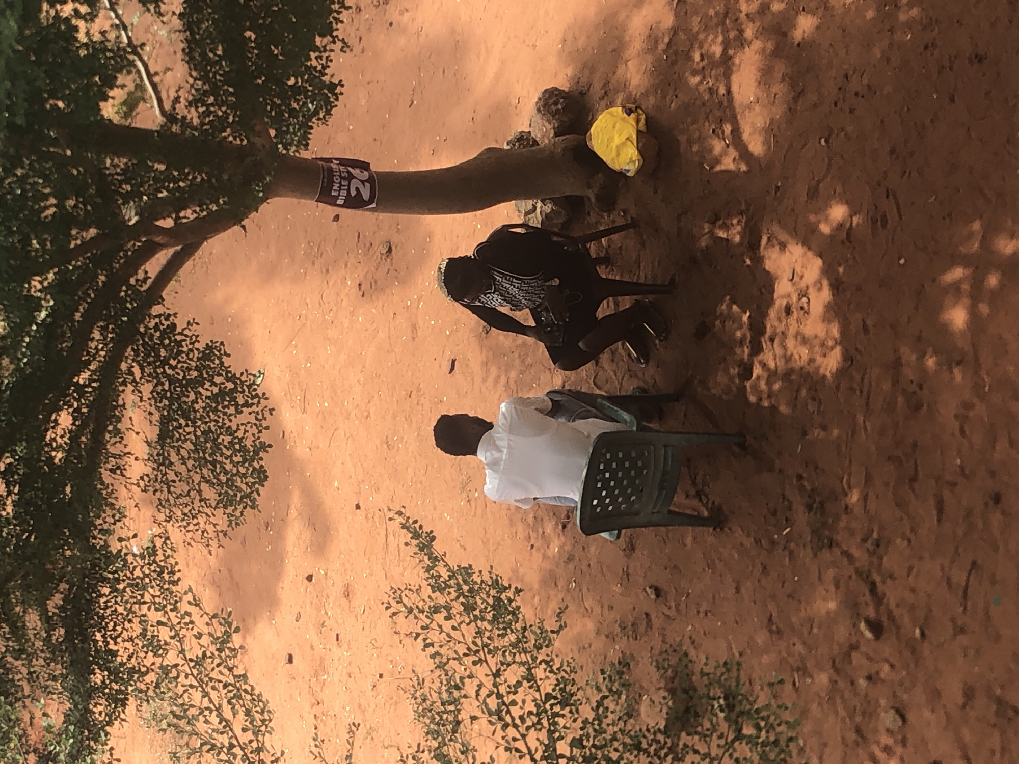 The scene is an outdoor setting in Nteje, Nigeria. The ground is reddish-brown soil, partially shaded by trees. Two people are present. One individual is seated in a green plastic chair, wearing a white shirt. Another individual is seated in a black chair, wearing dark clothing with a light-colored pattern on the upper garment. A yellow cloth lies near the base of a tree. The tree has a sign affixed to it, partially obscured, displaying the text "ENGLISH BIBLE ST".