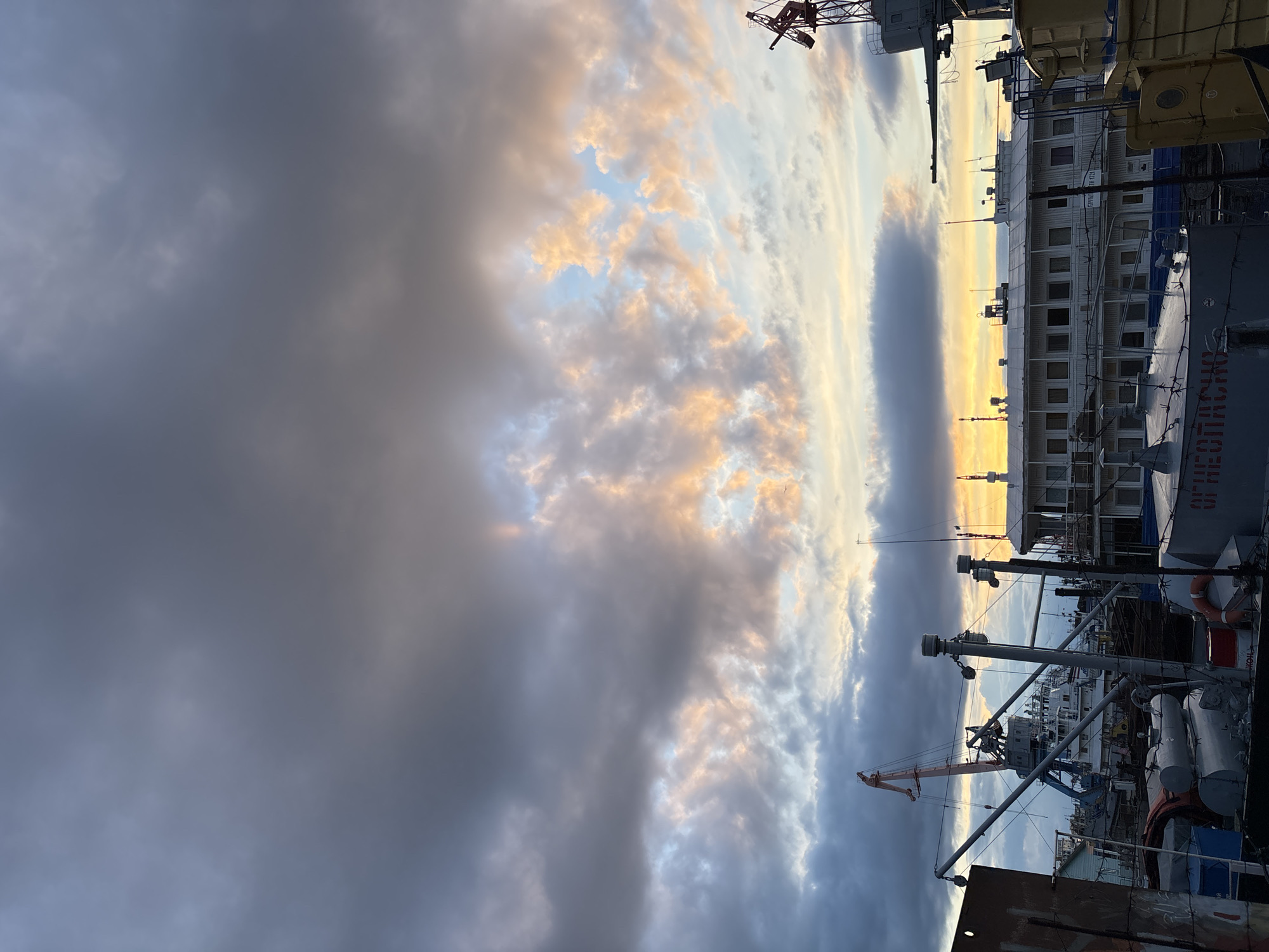 The image depicts a cloudy sunset sky over a port in what appears to be the city of Chaikovsky, Russia. Visible are elements such as port cranes, shipping containers, and various vessels. The cloudy sky contrasts with a strip of light near the horizon, suggesting the sun setting. The viewpoint is at ground level looking upward, and the orientation is sideways. The overall ambiance appears industrial.