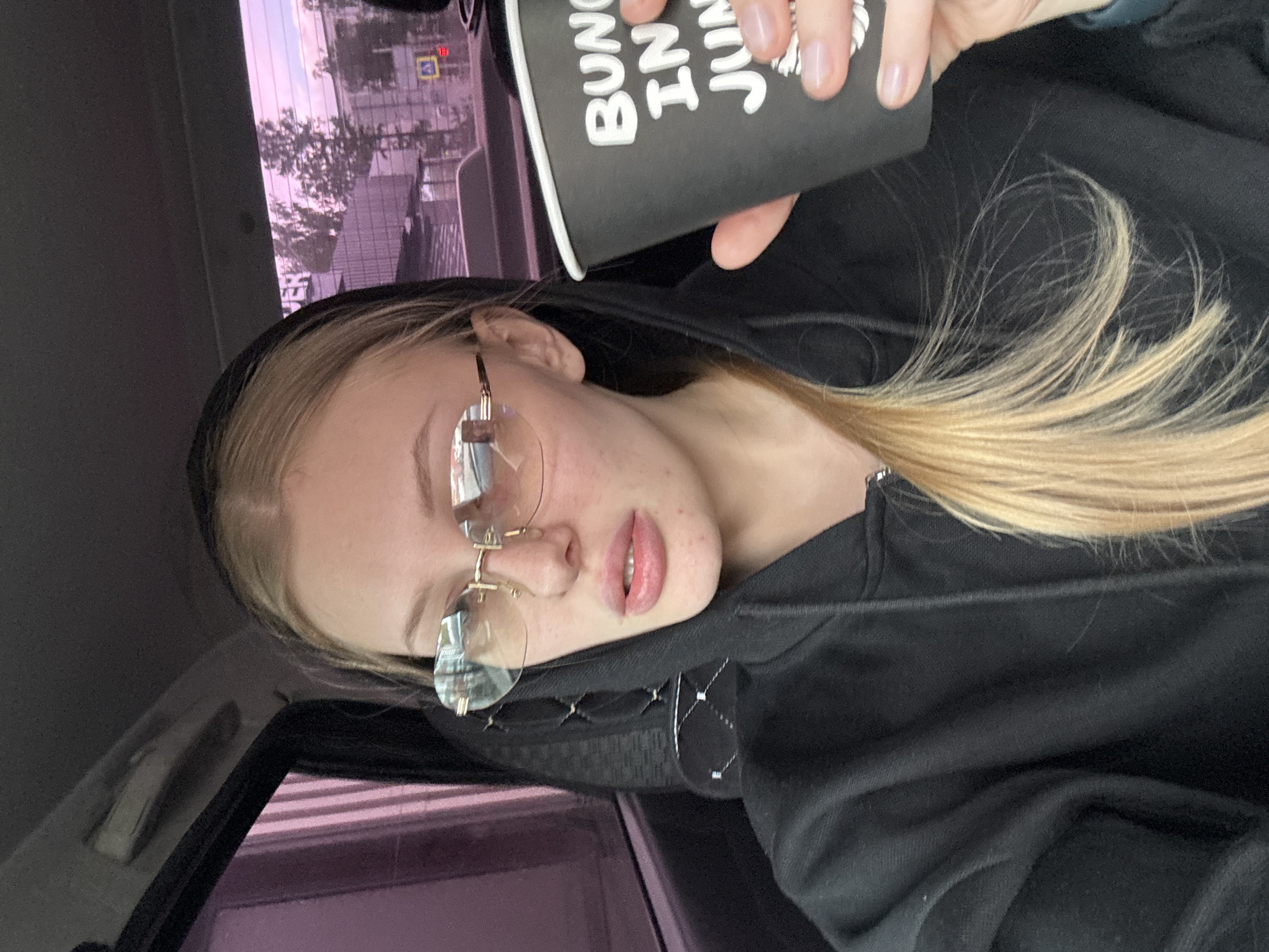 A young, light-skinned woman with blonde hair and light-colored eyes wearing rimless eyeglasses is holding a black coffee cup with white writing in a car. She is wearing a black hooded sweatshirt. The woman's location is evidenced by the view through the car window showing a building with Cyrillic writing and a street sign, suggesting a city location in Perm, Russia.