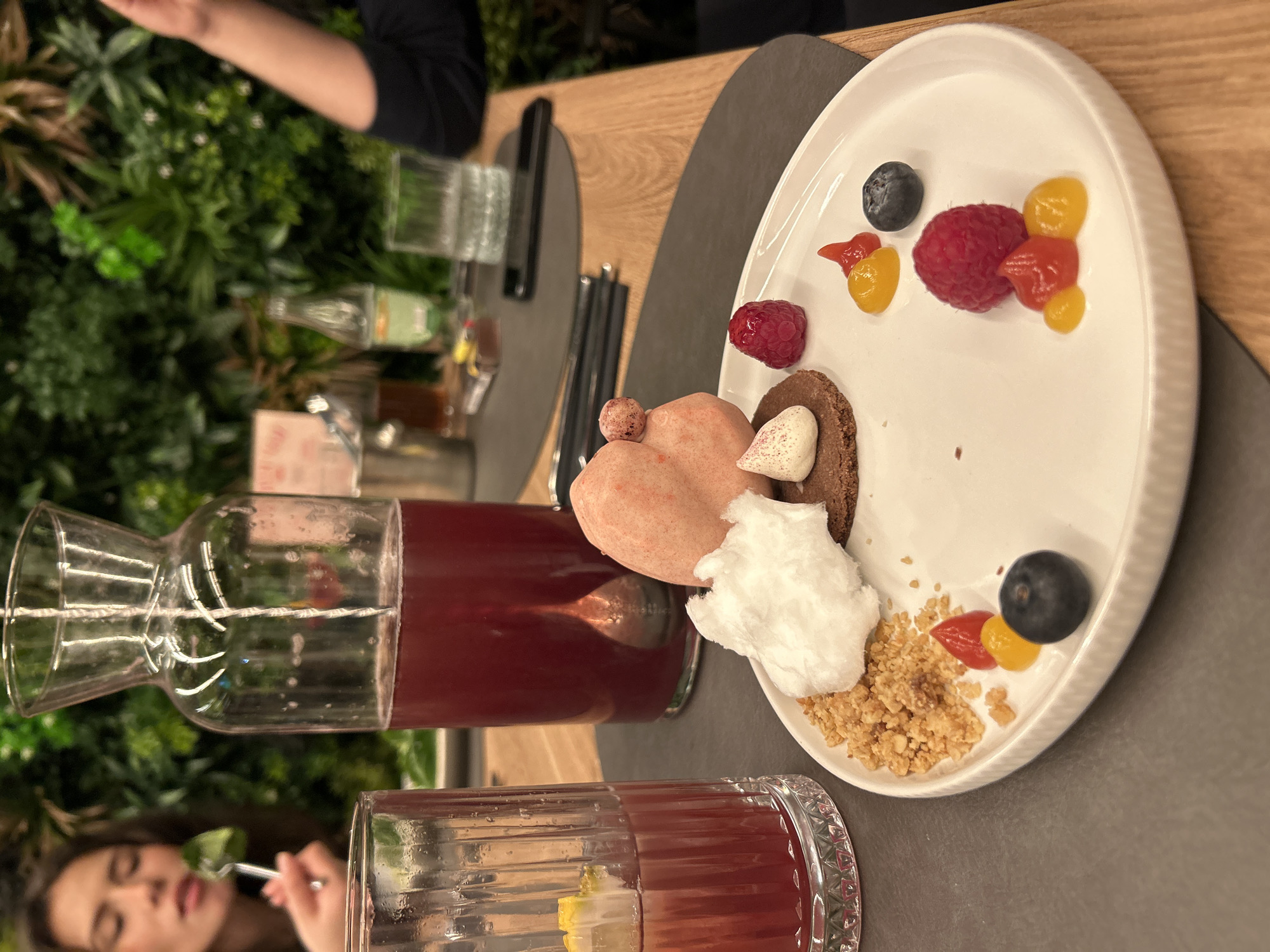 A close-up shot captures a dessert plate and drinks on a table, likely in a restaurant setting. The plate contains a variety of items including raspberries, blueberries, small colorful gel-like pieces, meringue, granola-like crumbles, and what appears to be a heart-shaped meringue cookie alongside a small chocolate cookie. To the left are glasses of a red-colored drink and a carafe filled with the same liquid. A woman is visible in the lower left corner eating from a glass. In the background, there are plants, suggesting either indoor decor or an outdoor terrace. The setting implies a dining experience, possibly in a restaurant or café. While Sofia, Bulgaria is mentioned in the prompt, there is no forensic information that supports the location being in Sofia, Bulgaria. Therefore, the location in the city of Sofia, Bulgaria is not possible to deduce from the content of the image.