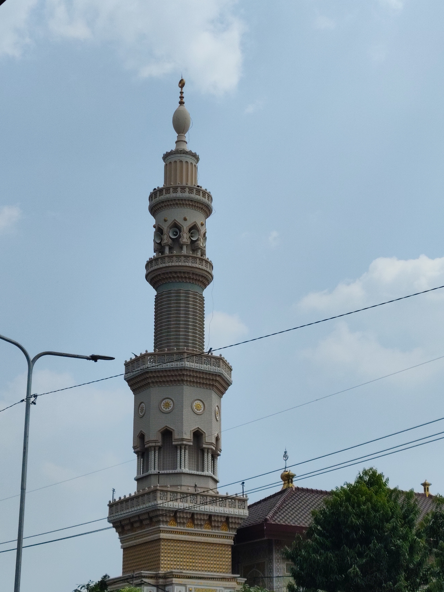 The image captures a tall, ornate minaret of a mosque in Dolopo, Indonesia, under a partly cloudy sky. The minaret is a light beige color with intricate decorative elements in gold and darker beige. It features multiple tiers with balconies, small windows, and speakers mounted on one of the upper sections. The top of the minaret is adorned with a finial topped by a crescent moon symbol. To the right of the minaret, the tiled roof of the mosque is visible, also topped with a smaller finial. Lush green trees partially obscure the lower part of the mosque and minaret on the right side of the frame. In the foreground, a dark lamp post with a modern design stands to the left, and several dark power lines crisscross the sky in front of the minaret and mosque. The scene appears to be a street-level view looking up at the mosque architecture.