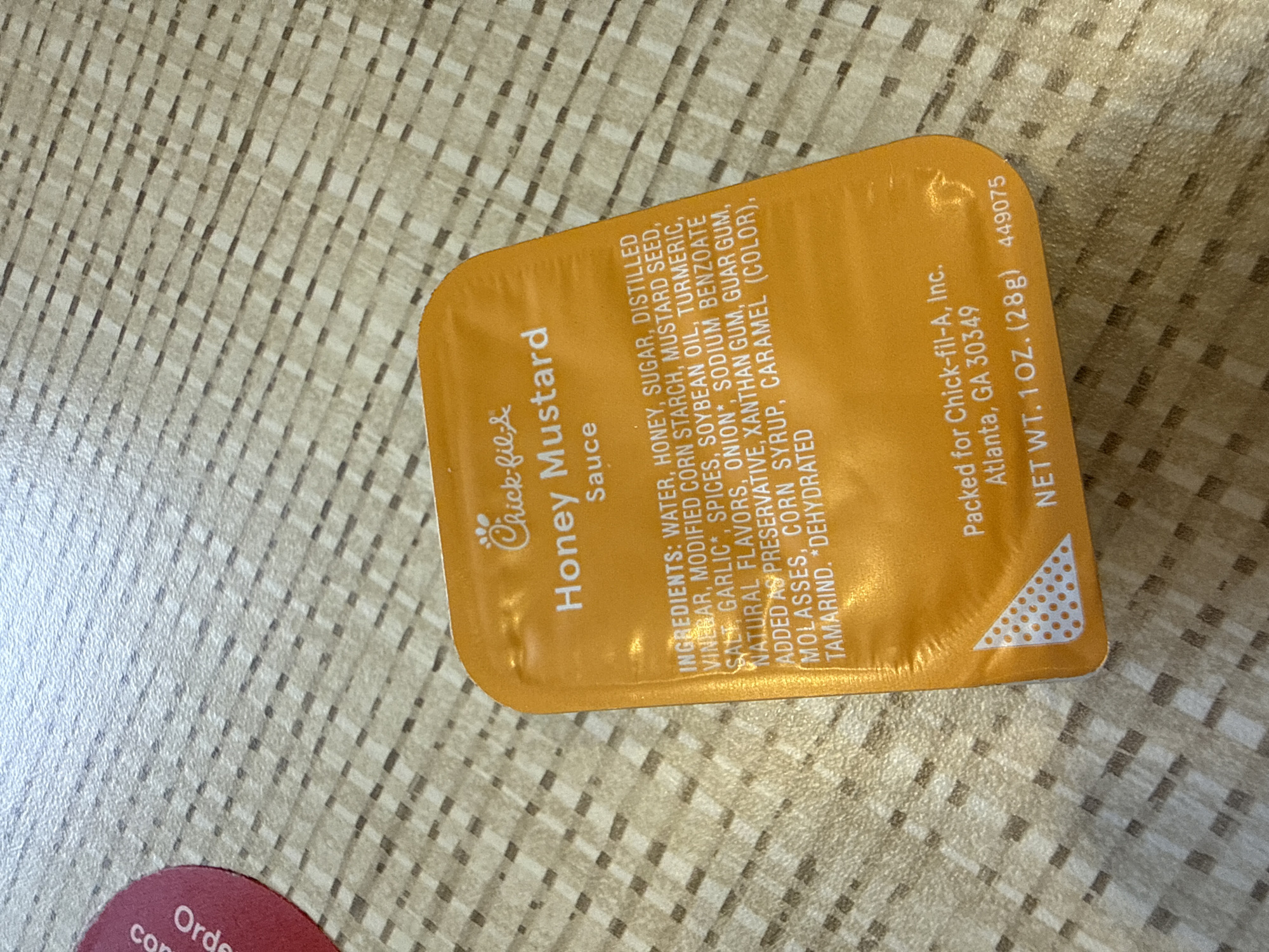 A close-up shot displays a packet of Chick-fil-A Honey Mustard sauce resting on a textured surface. The packet is predominantly yellow with the Chick-fil-A logo and "Honey Mustard Sauce" printed on its front. A list of ingredients is detailed along the side, including water, honey, sugar, distilled vinegar, and modified corn starch, among others. The bottom of the packet indicates it is packed for Chick-fil-A, Inc. in Atlanta, GA 30349, and states the net weight as 1 oz (28g). Part of a round item with the word "Order" is visible on the bottom left corner. The surface beneath the packet is a light beige with a repetitive textured pattern. The photograph offers a detailed view of the product packaging. It is not possible to verify the location is Stone Mountain.