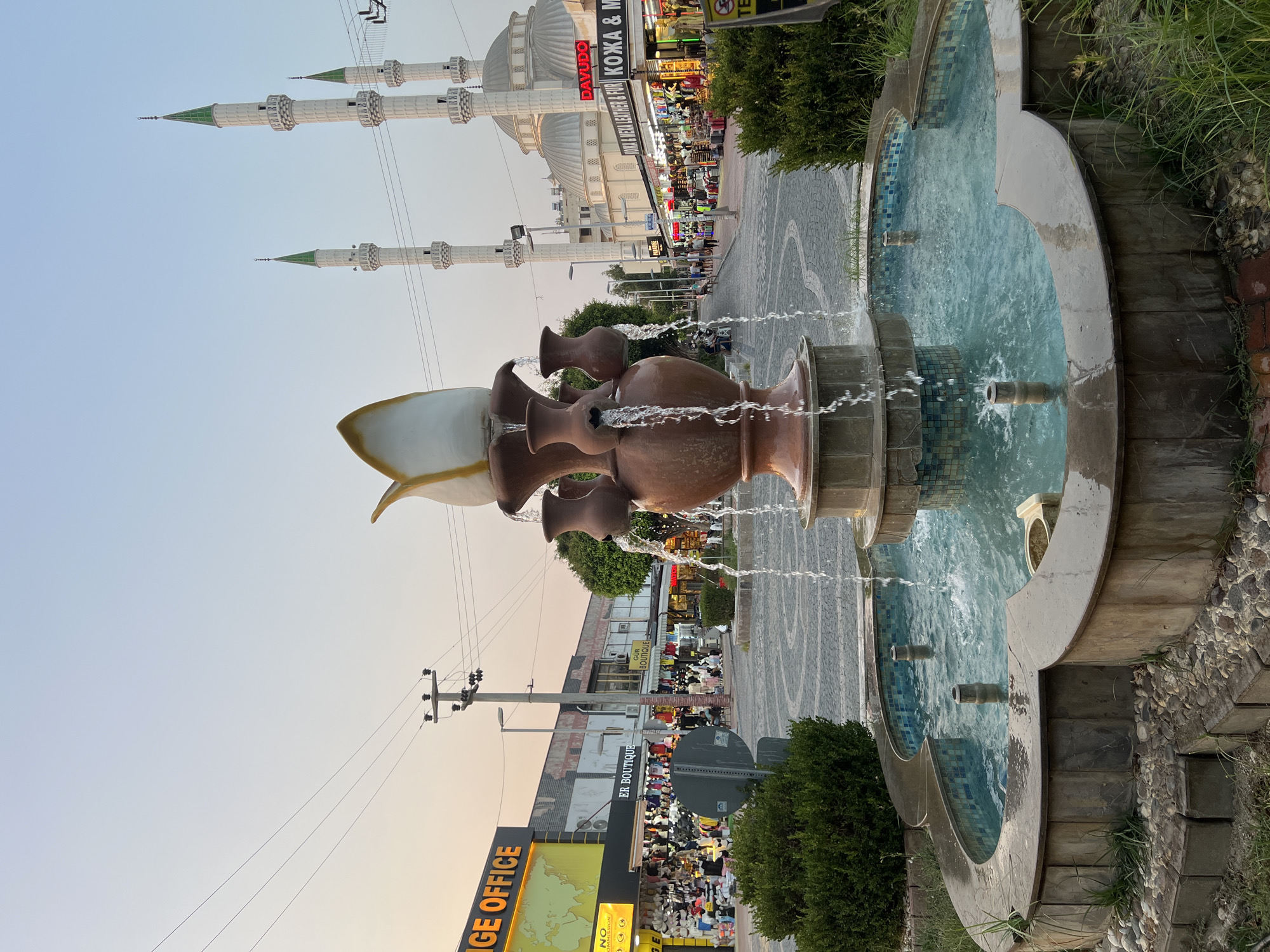 In an outdoor urban setting, a fountain with multiple terracotta-colored pots serves as the central foreground subject, with water cascading from its upper tiers into a tiled basin. Behind the fountain, a paved walkway is lined with shops on both sides. To the right, a large mosque with white minarets and domes dominates the background. Several people are visible walking along the street and browsing in the shops. Power lines and utility poles are present in the midground. The sky is clear and shows the soft light of either early morning or late evening. The overall scene suggests a bustling commercial area adjacent to a religious landmark.