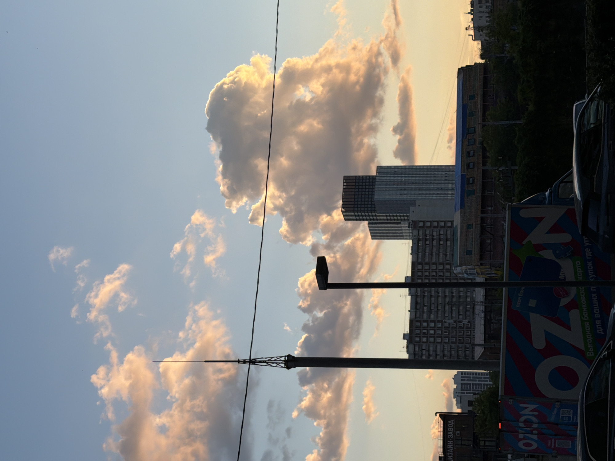 The image presents an urban landscape captured during what appears to be late afternoon or early evening, characterized by warm, golden light illuminating the clouds. The scene is set in Khimki, Russia.
The upper portion of the frame is dominated by a expansive sky, displaying a gradient from light blue to a soft, golden-orange hue near the horizon. Numerous cumulus clouds are scattered across the sky, with their edges and upper surfaces brightly lit by the low sun, creating a striking contrast with their shadowed undersides. A thin, dark power line stretches diagonally from the top left towards the bottom right of the sky, cutting across some of the cloud formations.
Below the sky, the horizon is filled with an array of urban structures. A tall, slender antenna-like mast with a distinctive latticework base stands prominently towards the left-center, alongside a dark street light pole with a rectangular fixture. Various multi-story buildings, ranging from older, blocky residential or commercial structures to more modern buildings with reflective facades, line the middle ground. Patches of dark green foliage from trees are visible amidst the buildings.
At the street level, portions of vehicles can be seen, including the front of a dark car on the far right and the side of a large truck or van. This vehicle features a colorful advertisement.
Visible text on signage includes "ДИЗАЙН-ЗАВОД" (DESIGN-PLANT/FACTORY) on a building in the lower left. The side of the truck prominently displays the "OZON" logo, accompanied by Russian text reading "Выгодная банковская карта" (Advantageous bank card) and "для ваших покупок где уг" (for your purchases where you...). Another smaller "OZON" logo is also visible below the main one. The overall impression is a dynamic city scene under a beautiful, luminous sky.