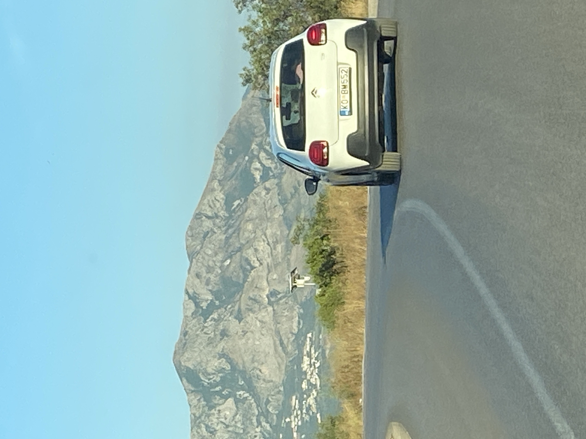 A white Citroen C3 is driving on a road in Dubraljevina, Montenegro. The car is viewed from behind, traveling on a paved road that curves to the left. In the background, a large mountain range dominates the horizon under a clear blue sky. Below the mountains and to the left of the car, a cluster of white buildings is visible, suggesting a settlement. Dry, yellowish vegetation lines the side of the road. A small, white, box-like structure with a solar panel on top stands on a pole to the left of the road, in front of the distant buildings. The car's license plate is visible, reading "KO BM552". The interior of the car shows a person with light-colored hair in the driver's seat and another person with dark hair in the passenger seat.
