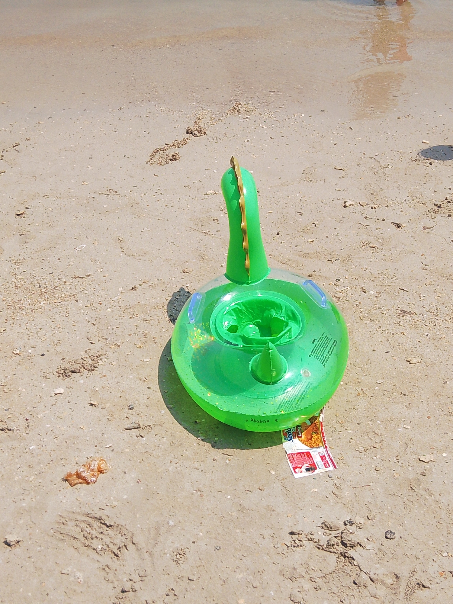 A green inflatable dinosaur-shaped baby float is positioned on a sandy beach near the water's edge in Pejagan, Indonesia. The float is transparent with green glitter inside and features a dinosaur head and tail. A piece of red and white packaging is tucked under the float. The sand is damp and shows ripples from receding waves. In the background, shallow water reflects the light. The scene is captured in daylight, with a shadow cast by the float.