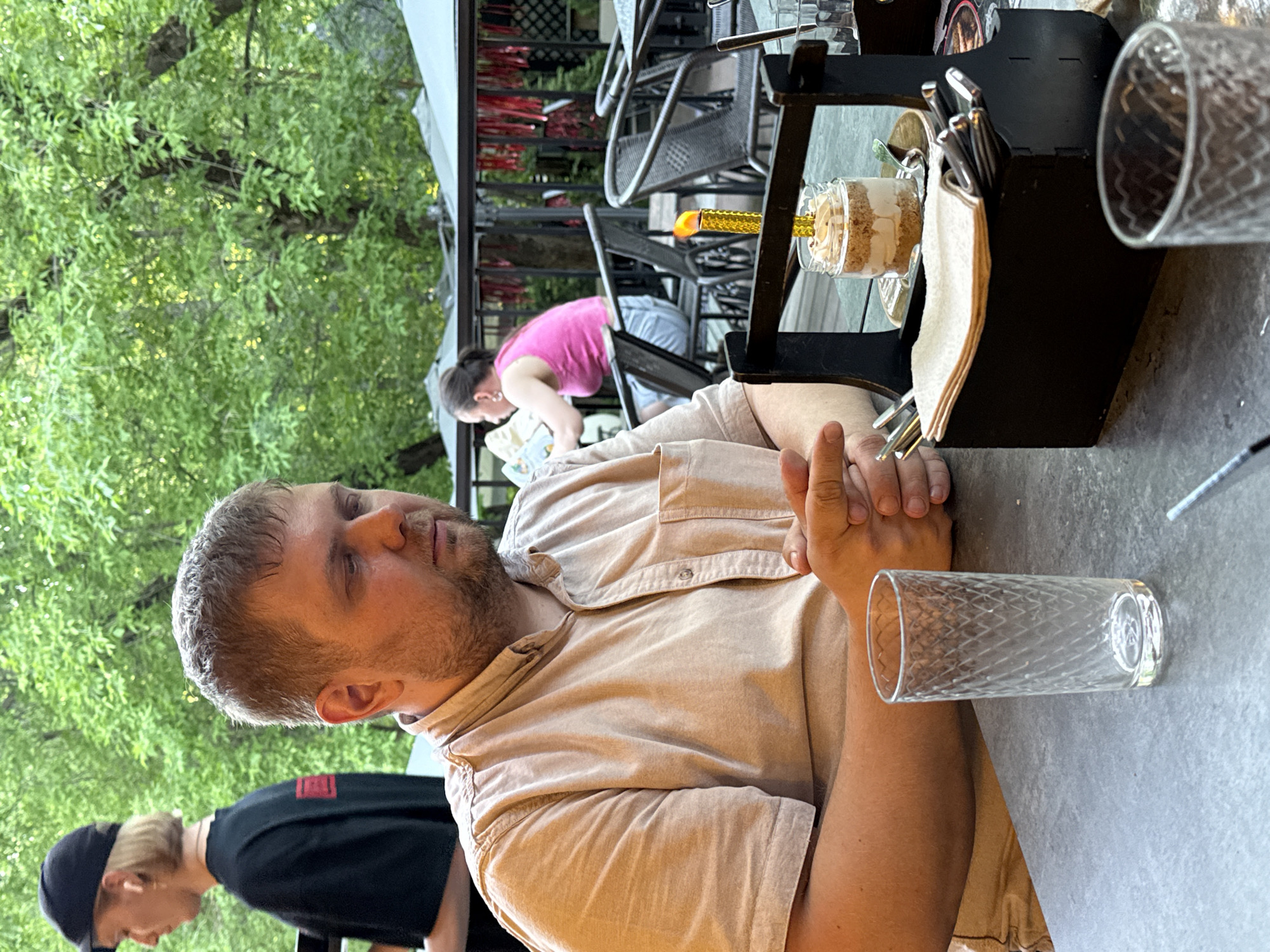A photo taken outside at a restaurant or cafe shows a man sitting at a table with another person standing next to him, a woman at a table behind him, trees to the side, a black condiment holder and dessert on the table, and a glass on the table. The man is light-skinned with short brown hair and a beard. He is wearing a tan collared shirt and has his hands clasped together. The person next to him is wearing a black shirt and a black baseball cap. The woman in the background is wearing a pink shirt and leaning over at a table. The table in the foreground is gray. The condiment holder is black and includes silverware and napkins.