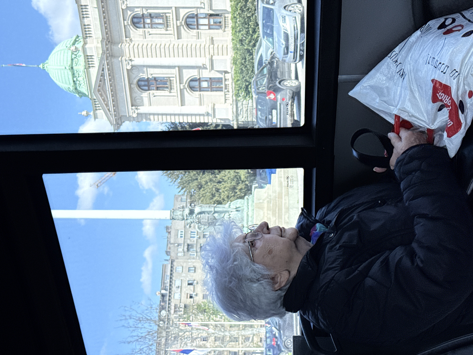 The image shows an elderly woman sitting in a vehicle (possibly a bus) looking out the window. She has short, gray hair and glasses. She is wearing a dark-colored jacket and holding a black, round object. Next to her is a white plastic shopping bag with red and black markings. Through the vehicle's window, the cityscape of Belgrade, Serbia, is visible. The National Assembly of Serbia can be seen along with a monument and several flags. Also visible are parked cars and buildings. The sky is blue with some clouds.
