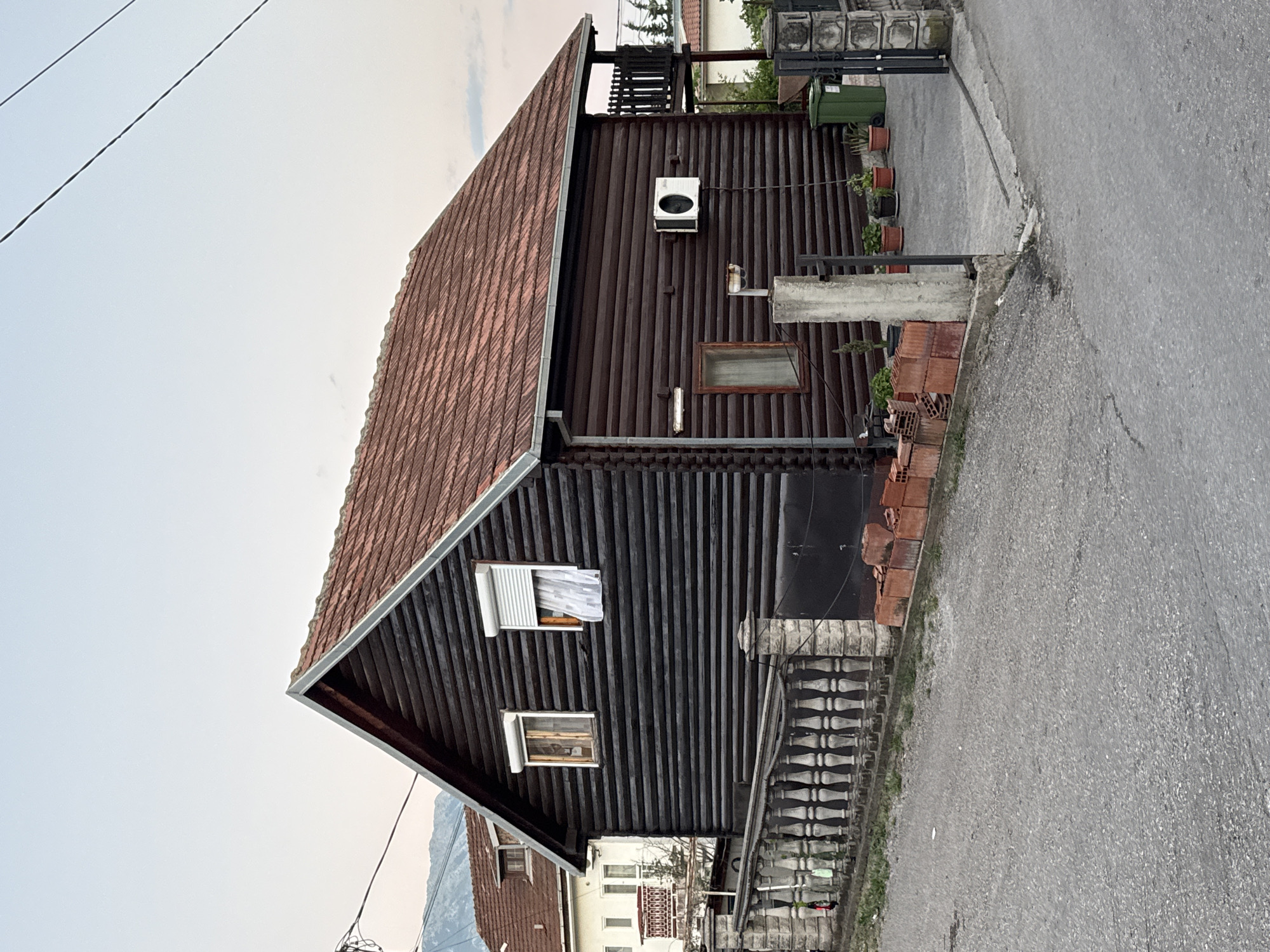 The image shows a dark brown log cabin-style house with a red tile roof, situated on a paved street in Burtaiši, Montenegro. A decorative stone fence runs along the front of the property. The house features several windows, including one with a white roller blind and sheer curtains. An outdoor air conditioning unit is mounted on the side of the house. To the right of the house, there is a small balcony with a railing and a green trash bin. Several potted plants are visible near the entrance and along the pathway. In the background, another building with a red tile roof and a balcony can be seen, along with trees and distant mountains under a pale sky. The scene appears to be a residential area, possibly in the late afternoon or early evening.