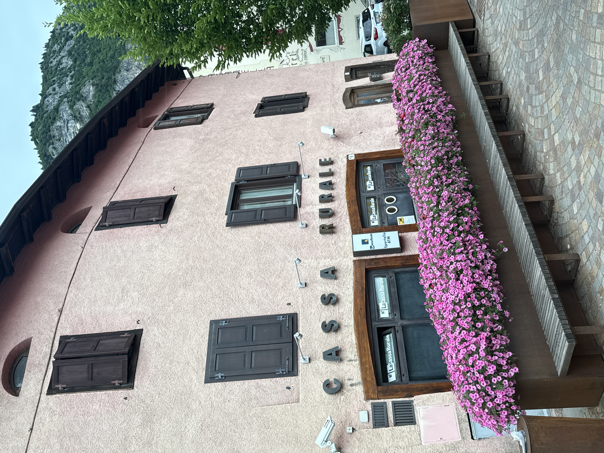 This is a medium-shot, eye-level, outdoor photograph of a building facade in Fai della Paganella, Italy. The building is painted a light pink and has dark brown shutters on its windows. A sign above the ground floor windows reads "CASSA RURALE." The ground floor features two windows with signs that say "La Cassa Rurale" and one with "Sportello ATM." A row of pink flowers cascades over a planter box in front of the windows, and a wooden bench sits in front of the planter. To the right of the building, a white car is parked. A lush green tree stands on the right side of the frame, and a mountainous landscape is visible in the background. The sky is overcast.