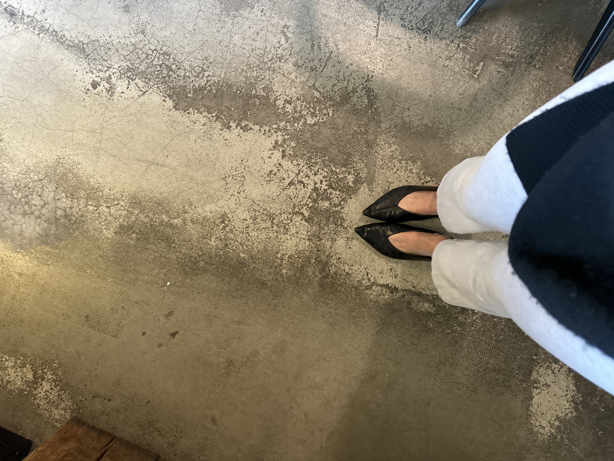 The scene depicts the lower extremities of an individual standing on a distressed concrete floor in an indoor environment, located in London, United Kingdom. The subject wears black, pointed-toe, flat-heeled shoes, light-colored (appearing white) loose-fitting trousers, and a black garment with a visible horizontal white segment across the upper thighs. The floor surface exhibits extensive wear, discoloration, and fine cracking, with irregular patches of light grey and darker brownish-grey, and subtle grid-like impressions in some areas. A dark, wooden structural element is partially visible in the upper-left corner of the frame. A metallic, silver-colored furniture component, consistent with a chair leg, is partially visible in the lower-right corner.