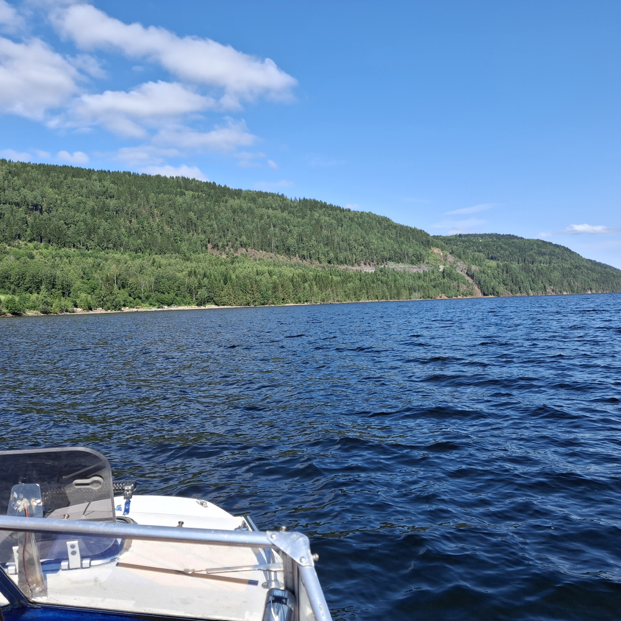This is a photograph taken from the perspective of a boat on a body of water, likely in Minnesund, Norway, given the context. The foreground shows the front of a white boat with a windshield and a metal railing. The water is a deep blue, with small waves indicating gentle movement. In the midground, a shoreline is visible, lined with trees and a road. The background is dominated by a large, densely forested hill that slopes down to the water's edge. The sky above is a clear blue with a few white, scattered clouds. The overall impression is a peaceful natural landscape during daylight.