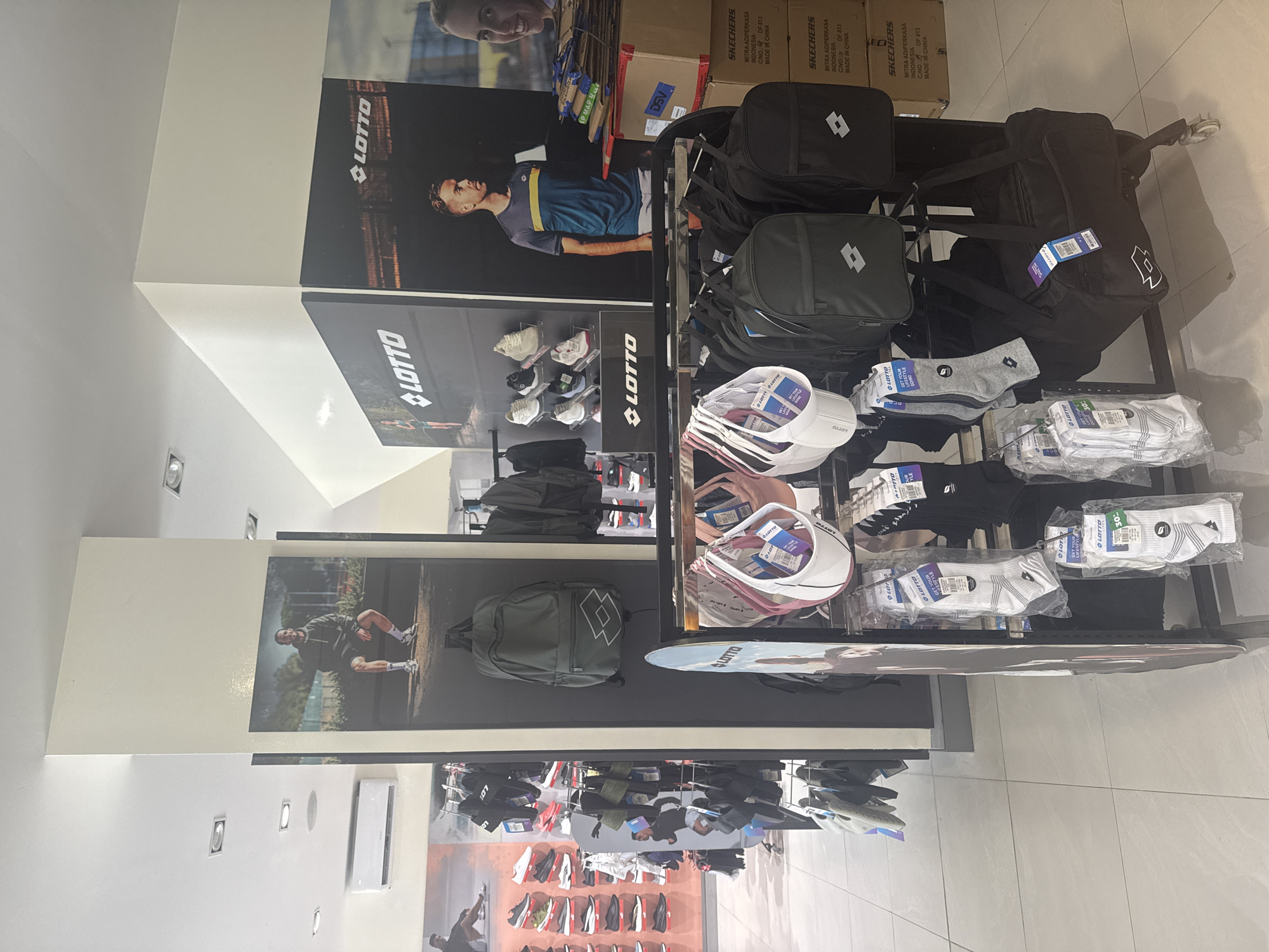 The image shows an indoor retail setting with LOTTO brand products. Displayed are bags, socks, and hats arranged on multi-tiered shelves. LOTTO advertisements are placed on the walls with images of sports figures. The location is likely a LOTTO store in North Kuta, Indonesia. Packaging of Skechers products is visible on the top shelf.
