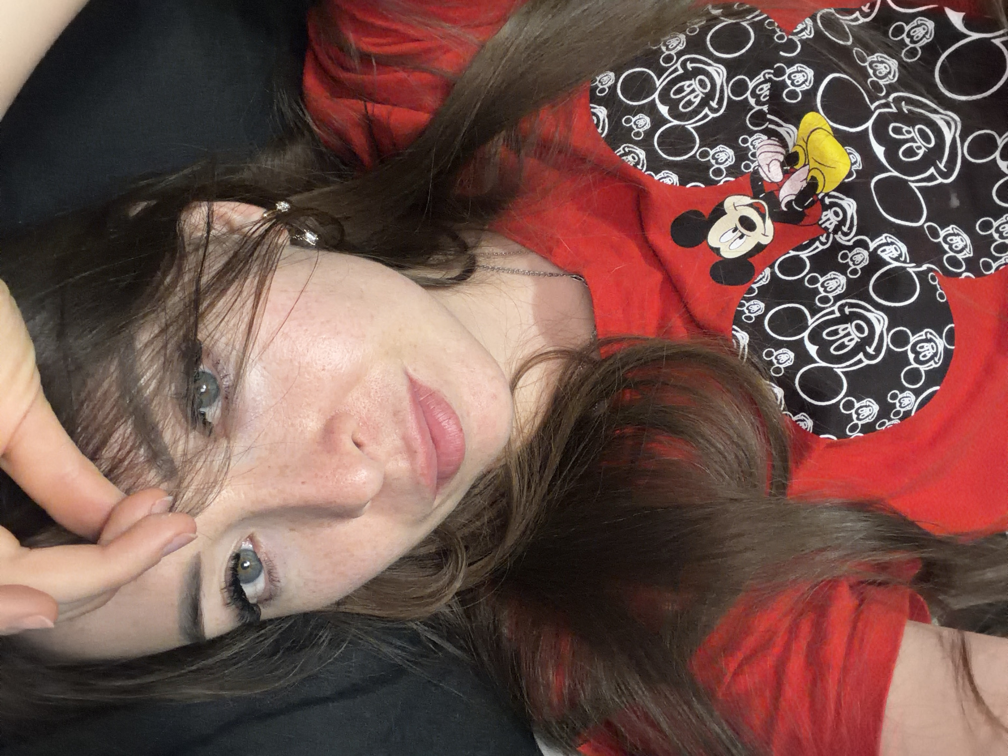 The media file is a close-up, high-angle shot of a young woman lying down and looking at the camera. She has long, dark brown hair with some reddish highlights visible, and fair skin with freckles. Her eyes are blue-green, and she has on makeup, including mascara and possibly eyeliner. Her lips are slightly parted and appear to have lipstick or a tinted balm on them. She is wearing a red t-shirt with a black pattern of Mickey Mouse heads. A silver chain necklace is visible around her neck. Her right hand is touching her forehead and hair, obscuring part of her face. The background is dark, suggesting an indoor setting. The lighting is soft, and the overall mood appears relaxed. No visible text is present in the image. The location context is "Япрыково, Russia."