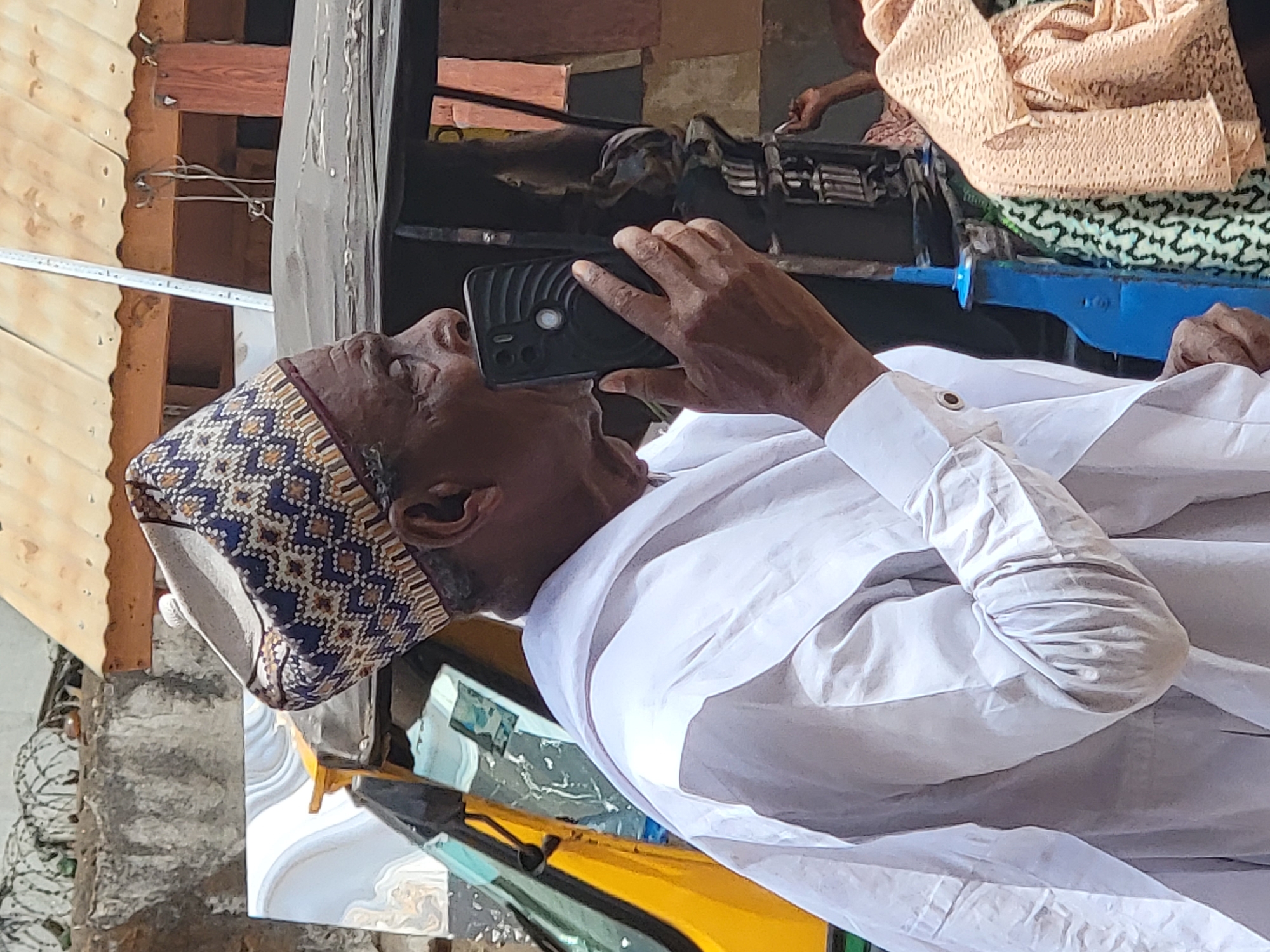 In Maiburiji, Nigeria, an elderly man wearing a patterned hat and a white robe holds a black smartphone to his ear. He is in a vehicle with a yellow exterior, possibly a taxi. In the background, there are other people, textiles, and what appears to be urban architecture.