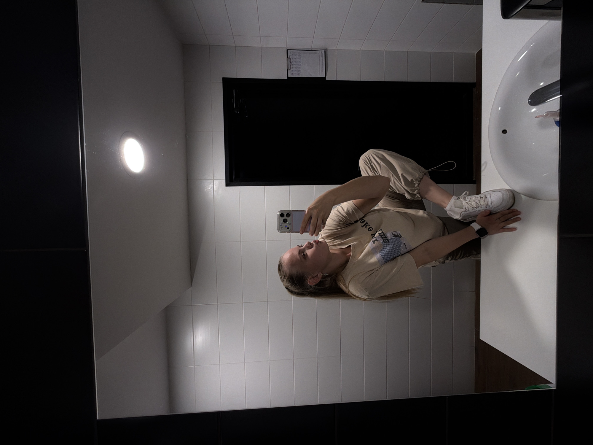 A young woman with blonde hair tied in a ponytail is taking a mirror selfie in a modern public restroom. She is wearing a light-colored, long-sleeved t-shirt and tan sweatpants. Her left leg is bent and raised, and she is holding a smartphone in her right hand, angled to capture the reflection. She is wearing white sneakers and a white smartwatch on her left wrist. The restroom features white tiled walls, a black door in the background, and a white sink with a faucet. The overall lighting is bright, with a ceiling light visible in the reflection. The environment suggests a clean and contemporary public facility. The image does not contain any visual cues that would allow for identification of the specific location within the city of Izhevsk, Russia.