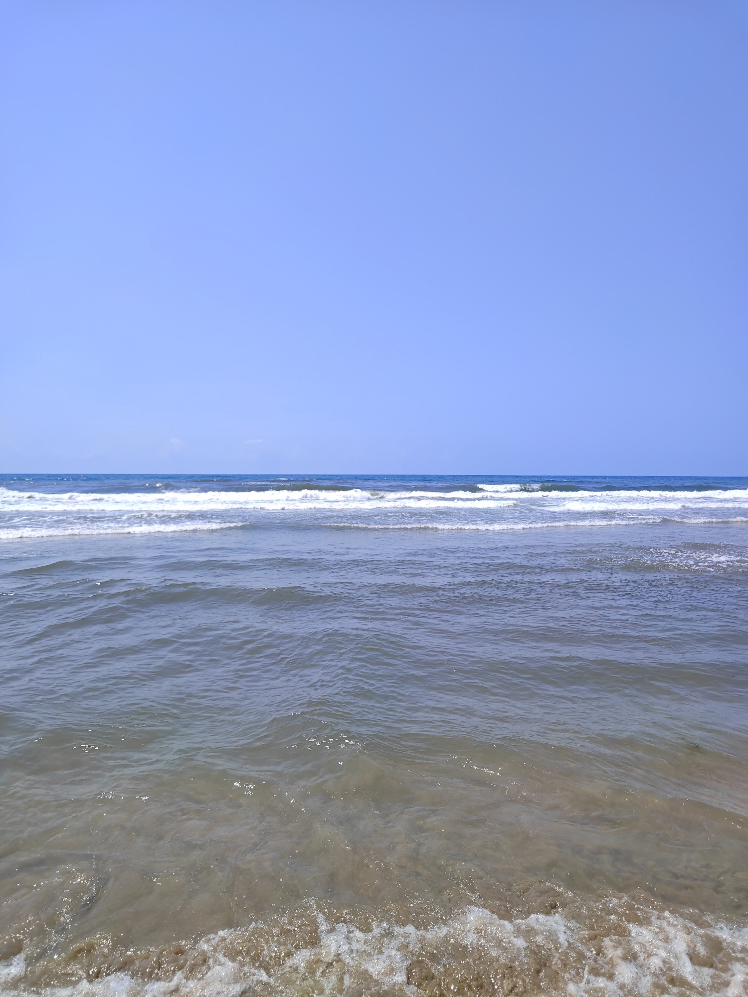 The image depicts a serene seascape on a clear, sunny day. The upper portion of the image is dominated by a vast expanse of pale blue sky, devoid of clouds. Below the sky, a thin, dark blue line marks the horizon where the sea meets the sky. Further down, multiple rows of gentle waves are visible, with white foam cresting on the outermost waves, indicating a receding tide. The water itself transitions from a deeper blue further out to a more translucent, sandy brown color closer to the shore, revealing the seabed beneath. The foreground is filled with shallow water and the sandy shore, where small waves are breaking with foamy edges. The scene suggests a tranquil beach environment, characteristic of coastal areas. This specific view likely corresponds to one of the beaches along the coastline of Pietrasanta, Italy, which is renowned for its sandy shores and clear waters. The image captures a moment of natural beauty and calm.