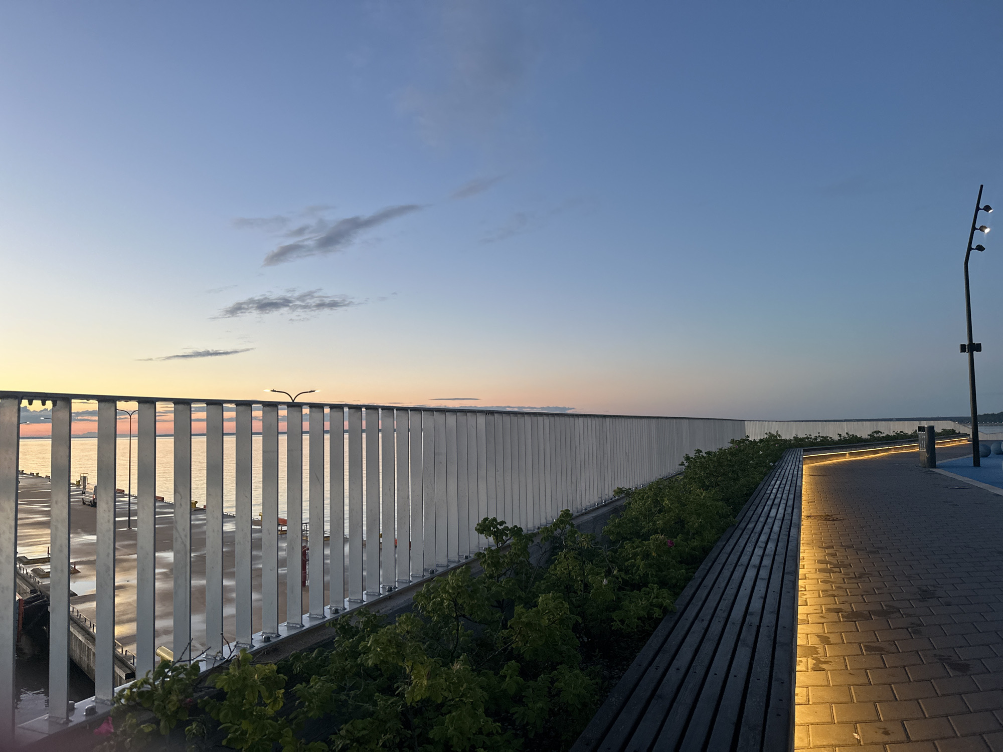 The image depicts a waterfront promenade at dusk in Tallinn, Estonia, likely in the area of the Old City Harbor or Seaplane Harbor. The sky is a gradient of soft blue and pale orange, indicating the sun has recently set. A modern, white, vertical-slatted fence runs along the left side of the promenade, separating it from a harbor area. Beyond the fence, calm water reflects the sky's colors, and a pier with some docked boats and yellow bollards is visible.
To the right of the fence, a paved walkway made of grey interlocking stones runs parallel to the water. Integrated into the edge of a dark wooden bench bordering the walkway are subtle, warm LED lights that illuminate the paving stones and cast long shadows. Beyond the bench is a strip of lush green bushes and some flowering plants. Further along the promenade, a contemporary streetlamp with three lights stands against the darkening sky. The overall atmosphere is serene and modern. No people are visible in the scene.