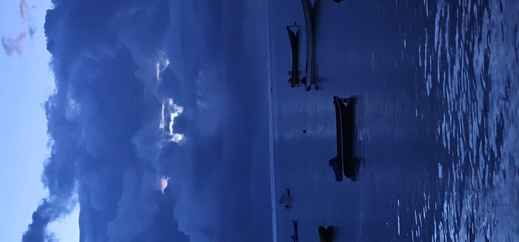 The image depicts a body of water with several boats in the foreground and a stormy sky in the background. The sky is predominantly dark blue, with thick, heavy clouds and patches of light peeking through. The boats are mostly small and appear to be moored or anchored. The water is dark and has a rippled texture, suggesting wind or current. The location is likely in Denpasar, Indonesia.